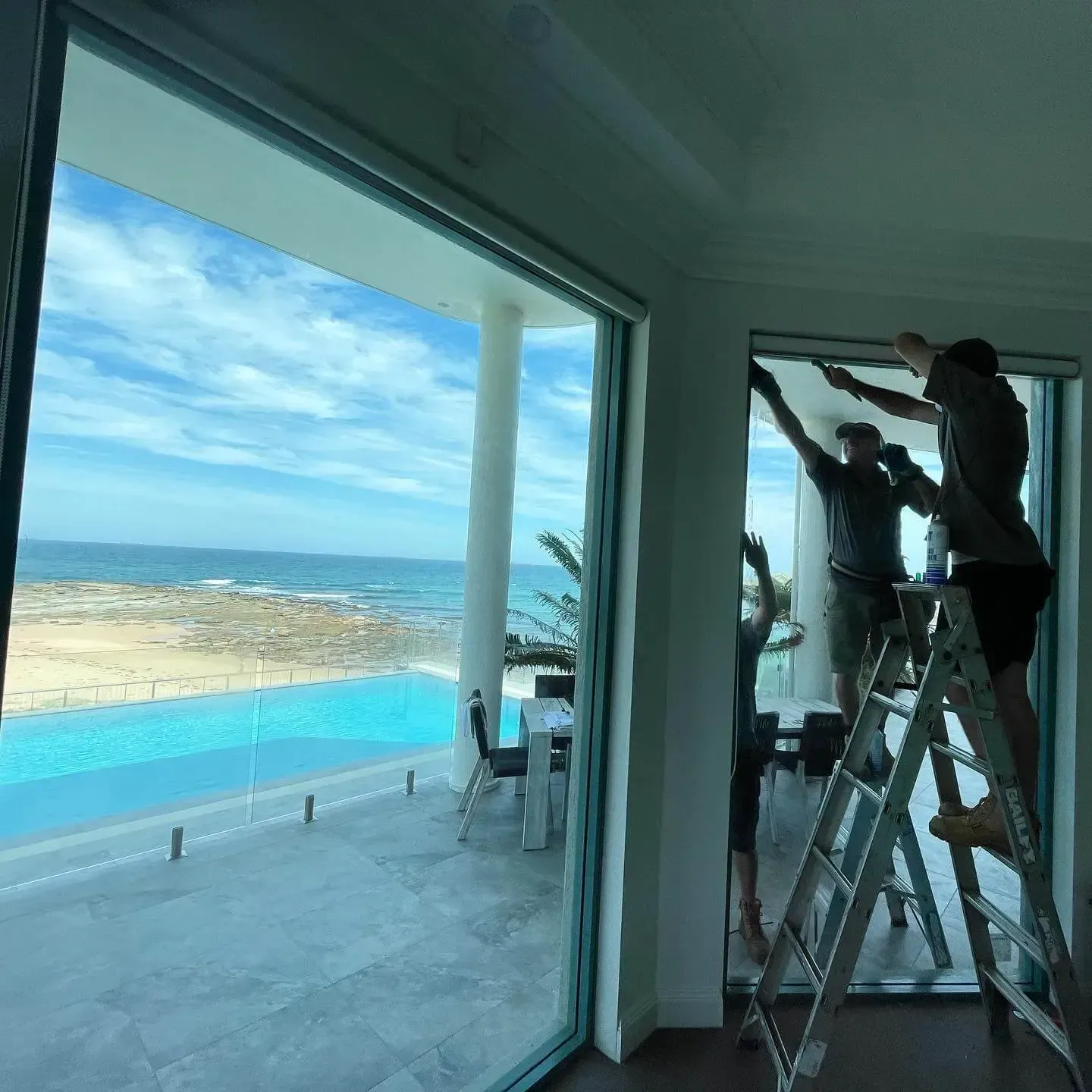 Two Men Are Standing on A Ladder in Front of A Window with A View of The Pool  — Tuggerah Lakes Glass In Tumbi Umbi, NSW