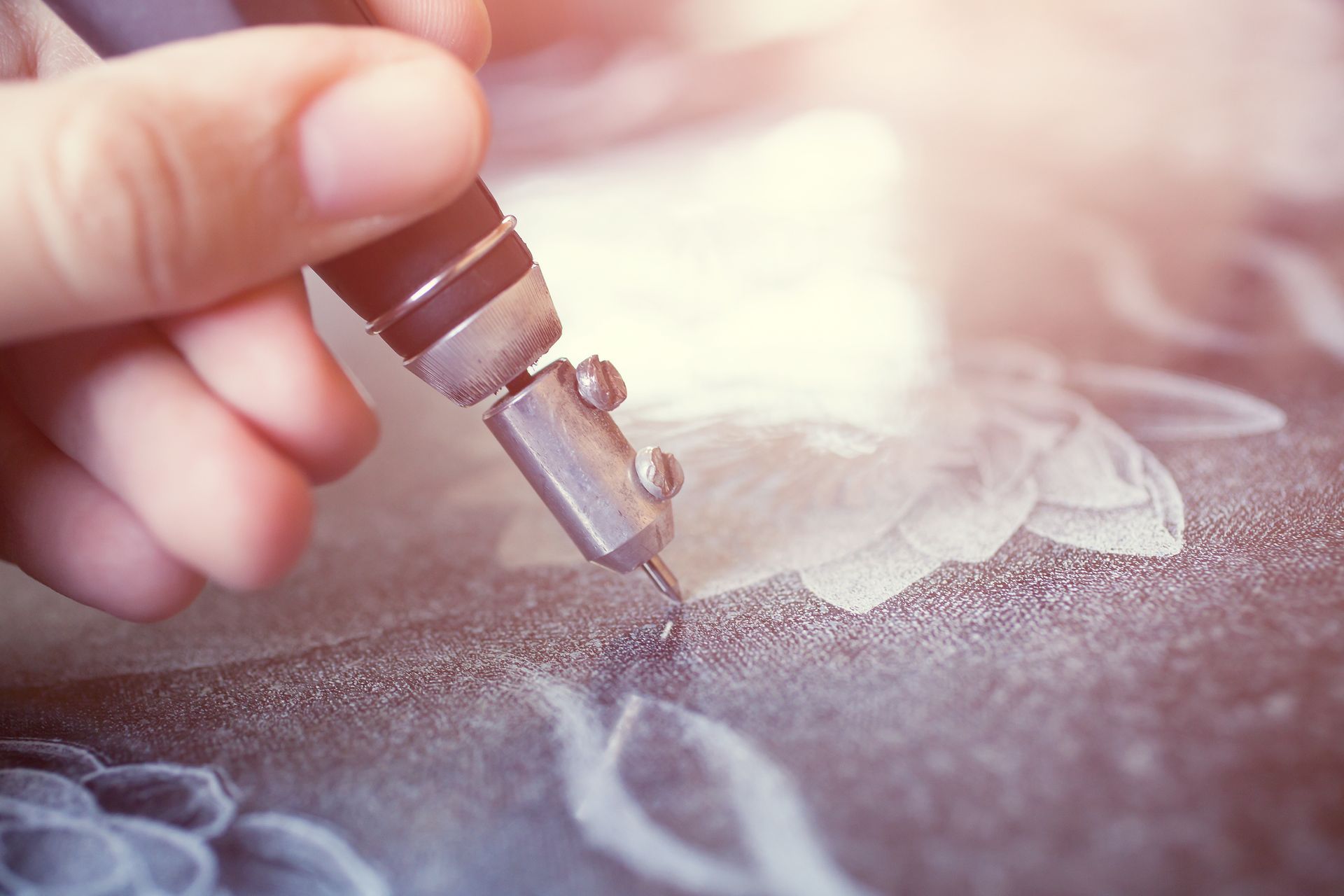 Hand using a glass cutter to etch a floral design on a dark surface, with bright lighting.