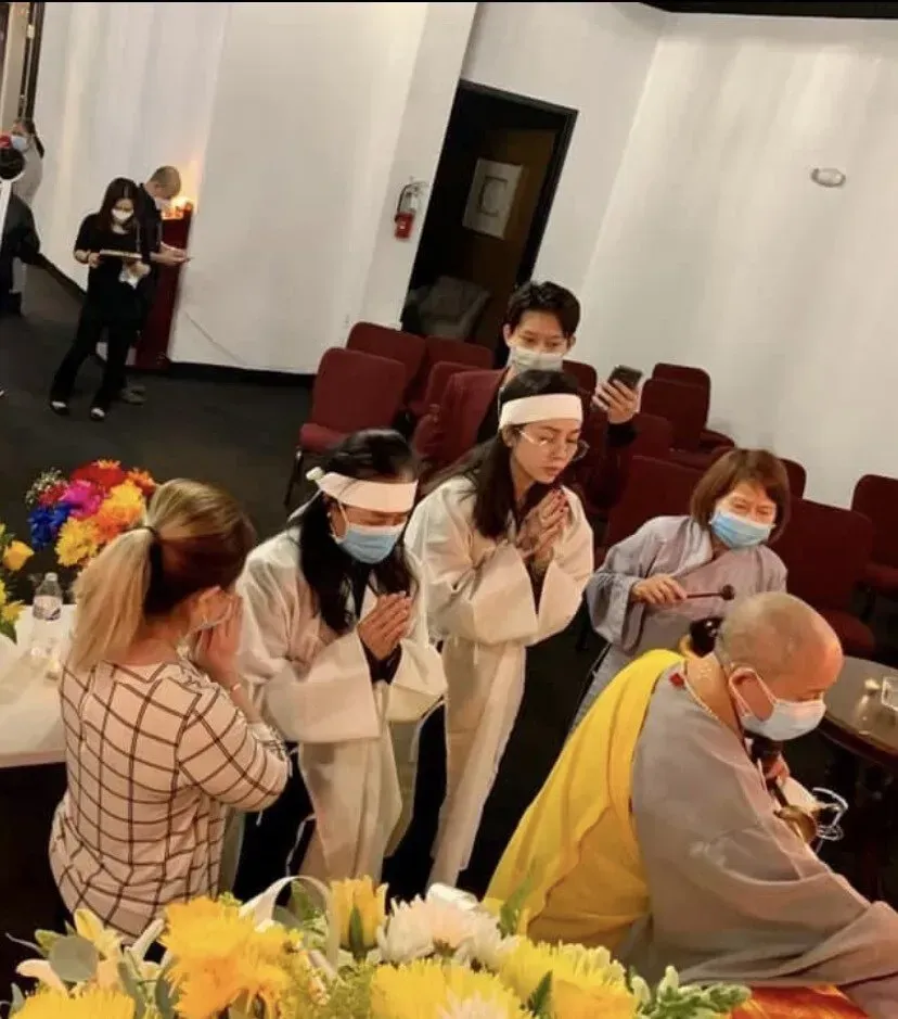 People gathered indoors, wearing face masks and robes, in a quiet room with red chairs and flowers.