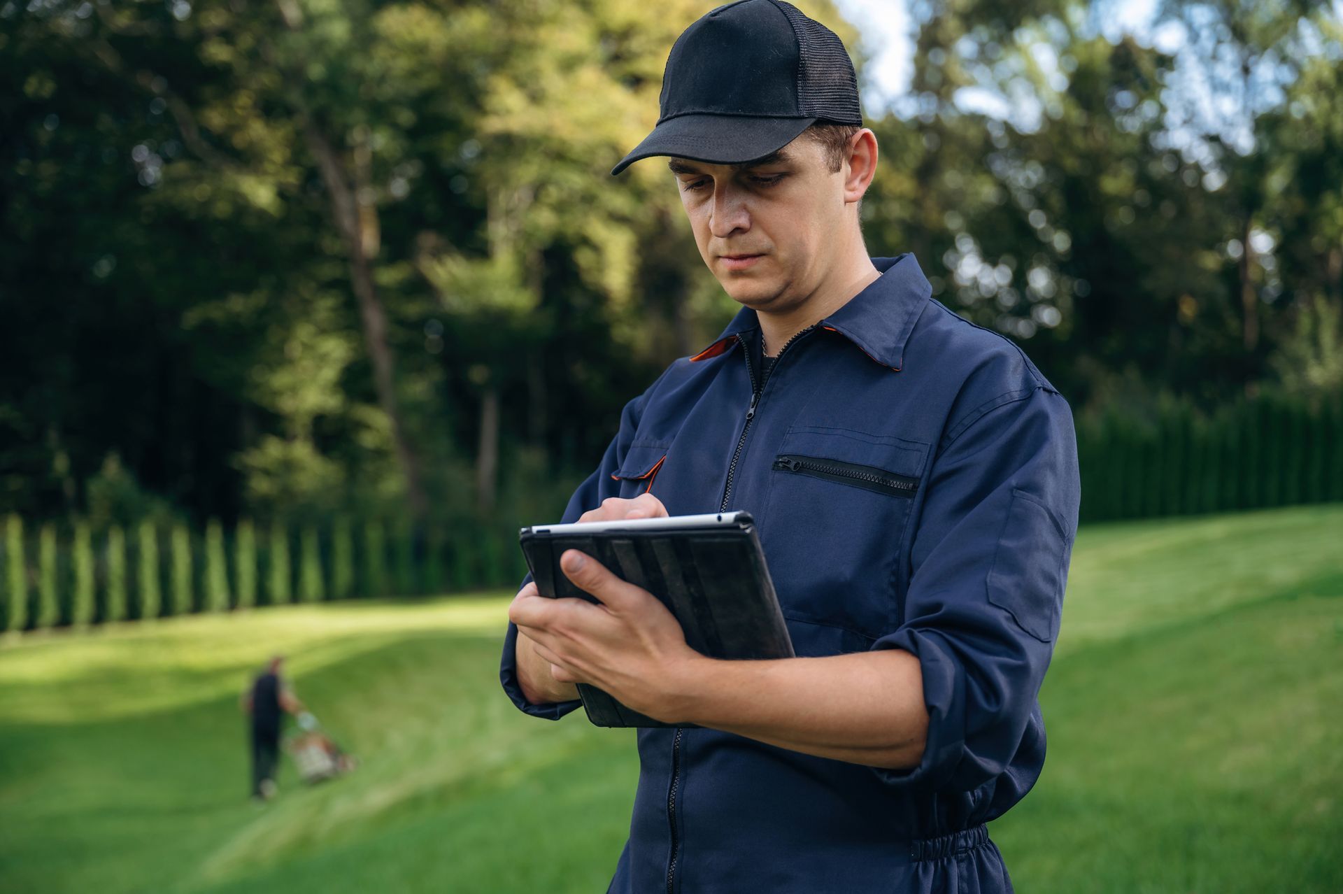 A professional in a blue uniform and black cap uses a tablet outdoors, with a person mowing a lawn in the background.