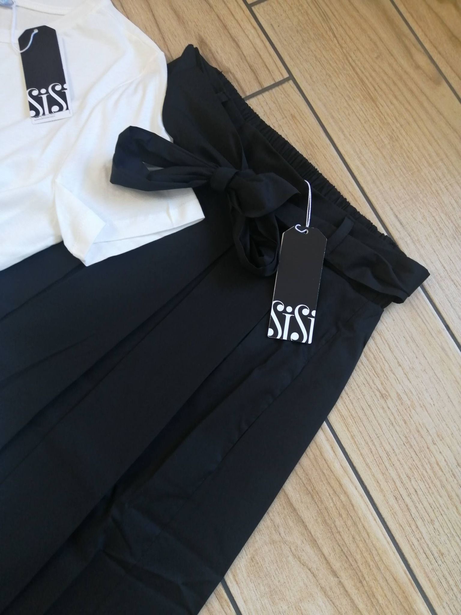 Una maglietta bianca e pantaloni neri con cintura in vita e cartellini del negozio attaccati, distesi su un pavimento di legno chiaro.