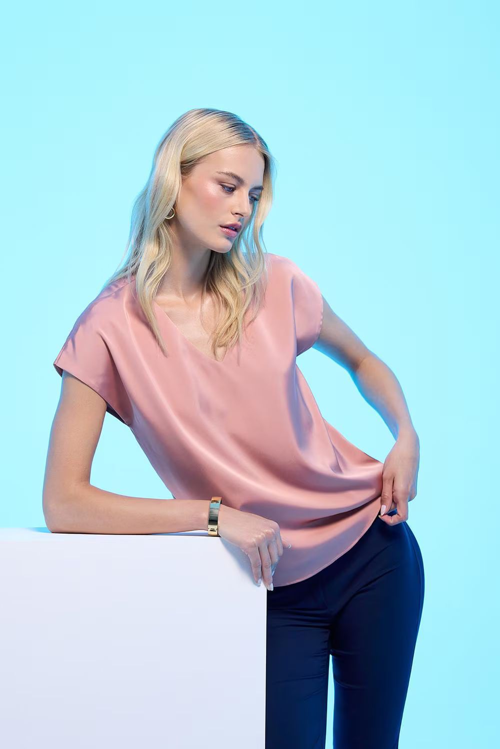 Una persona con i capelli biondi, che indossa una maglietta a maniche corte rosa cipria e pantaloni scuri, si appoggia a un blocco bianco contro una luce blu.