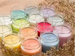 Una collezione di candele multicolori in barattoli di vetro, disposte insieme su una superficie rustica con elementi botanici essiccati.