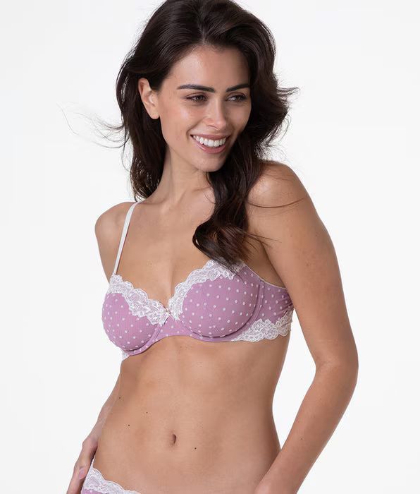 Persona sorridente che indossa un reggiseno a pois lilla chiaro con bordi in pizzo bianco su uno sfondo bianco uniforme.