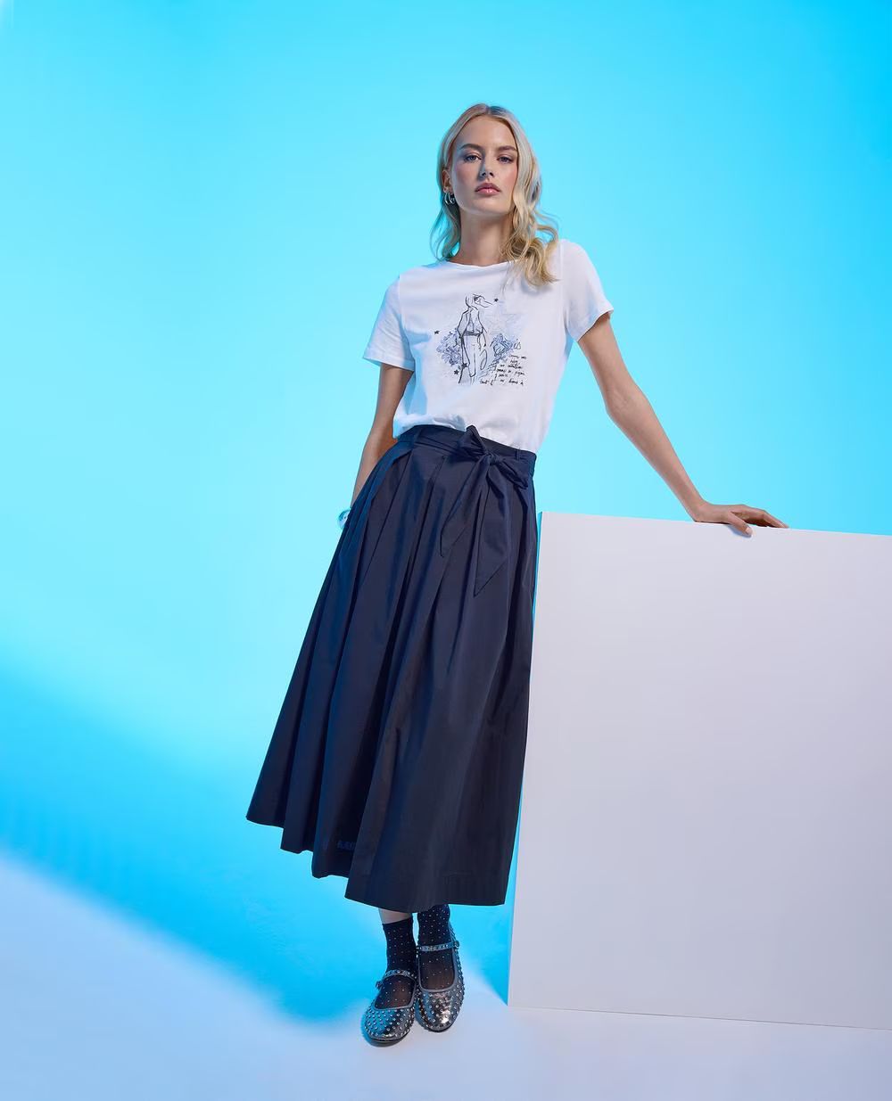 Una persona posa davanti a uno sfondo blu, indossando una t-shirt bianca con una stampa, una lunga gonna scura e ballerine argentate.