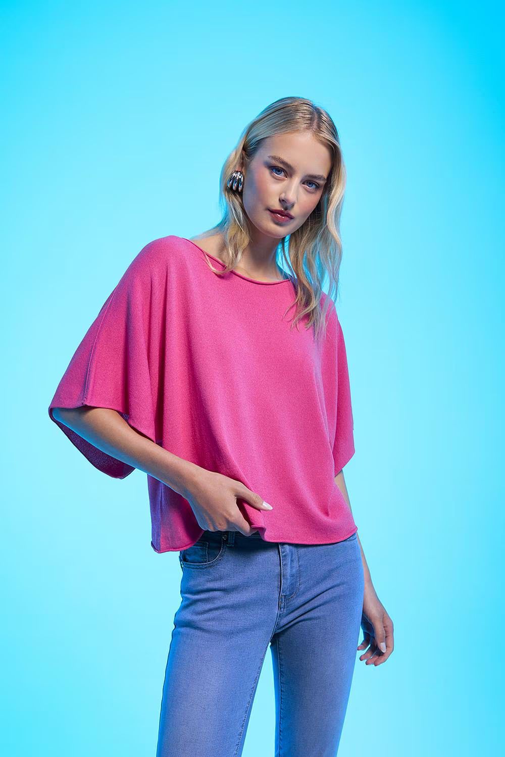 Una persona con lunghi capelli biondi indossa una maglietta rosa acceso a maniche corte e jeans blu su uno sfondo blu brillante.