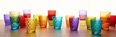 Una varietà di bicchieri di vetro colorati e dalle diverse texture, disposti su una superficie di legno su uno sfondo bianco.