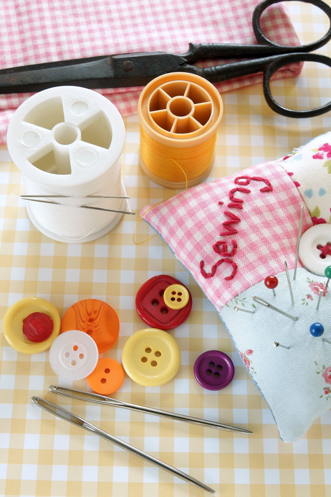 Una collezione di accessori per il cucito, tra cui rocchetti di filo, aghi, bottoni colorati e un puntaspilli su una superficie a quadri.