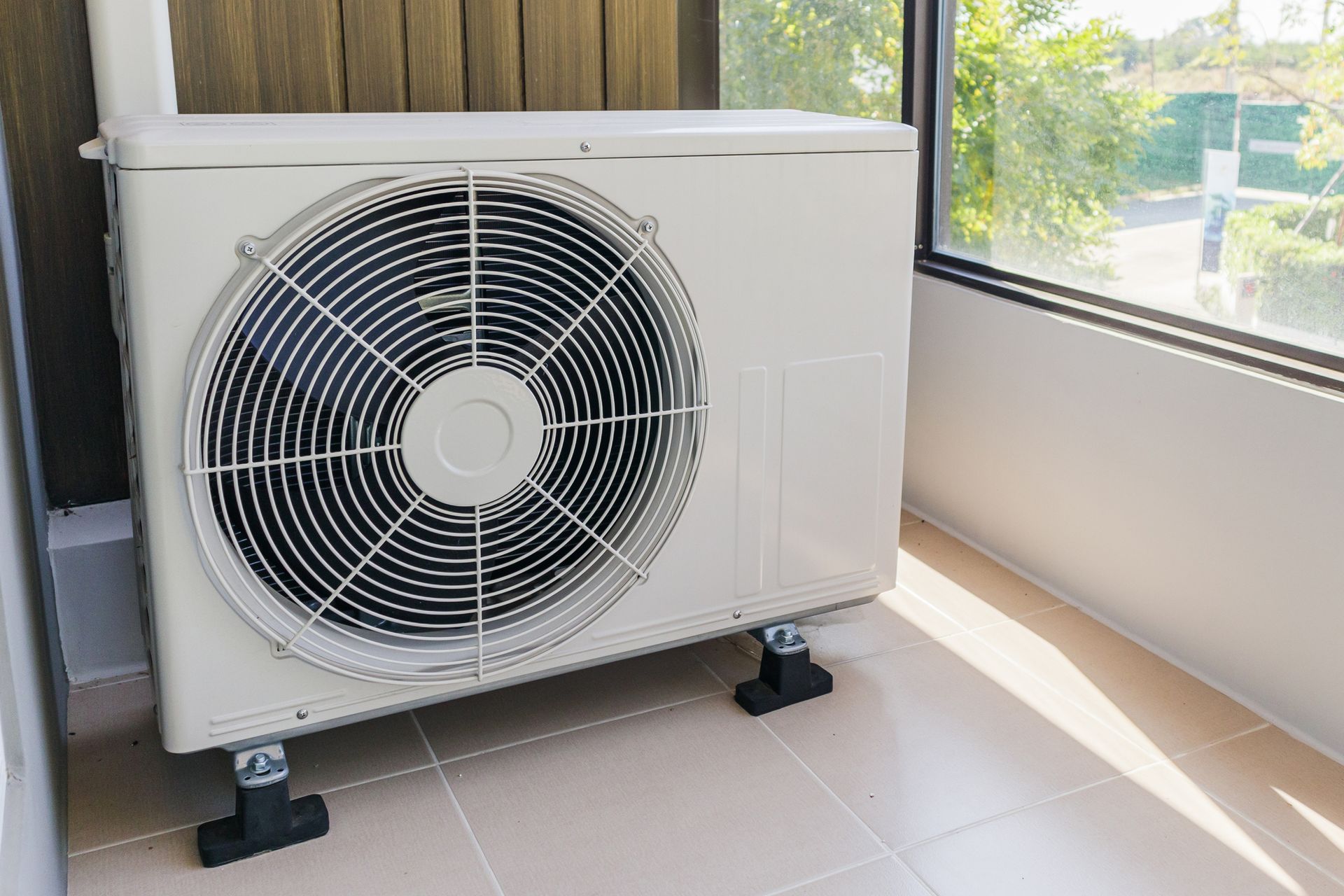 White air conditioning unit, with fan, sits on black blocks near a window.