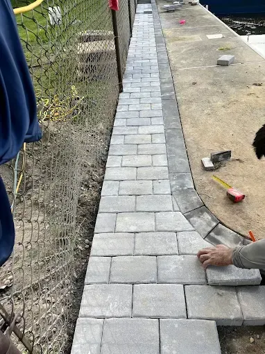 Person laying grey paving stones along a fence.