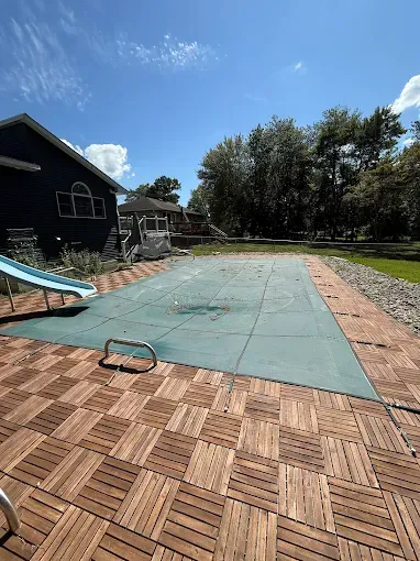 Pool covered with a blue tarp, wooden patio, a blue slide, and a blue house under a sunny sky.