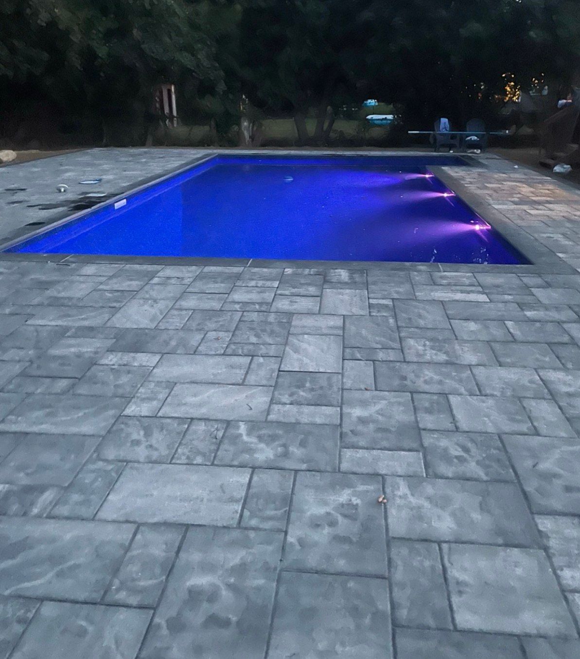 Green, algae-filled rectangular pool surrounded by brick patio, lush green trees in the background under a clear sky.