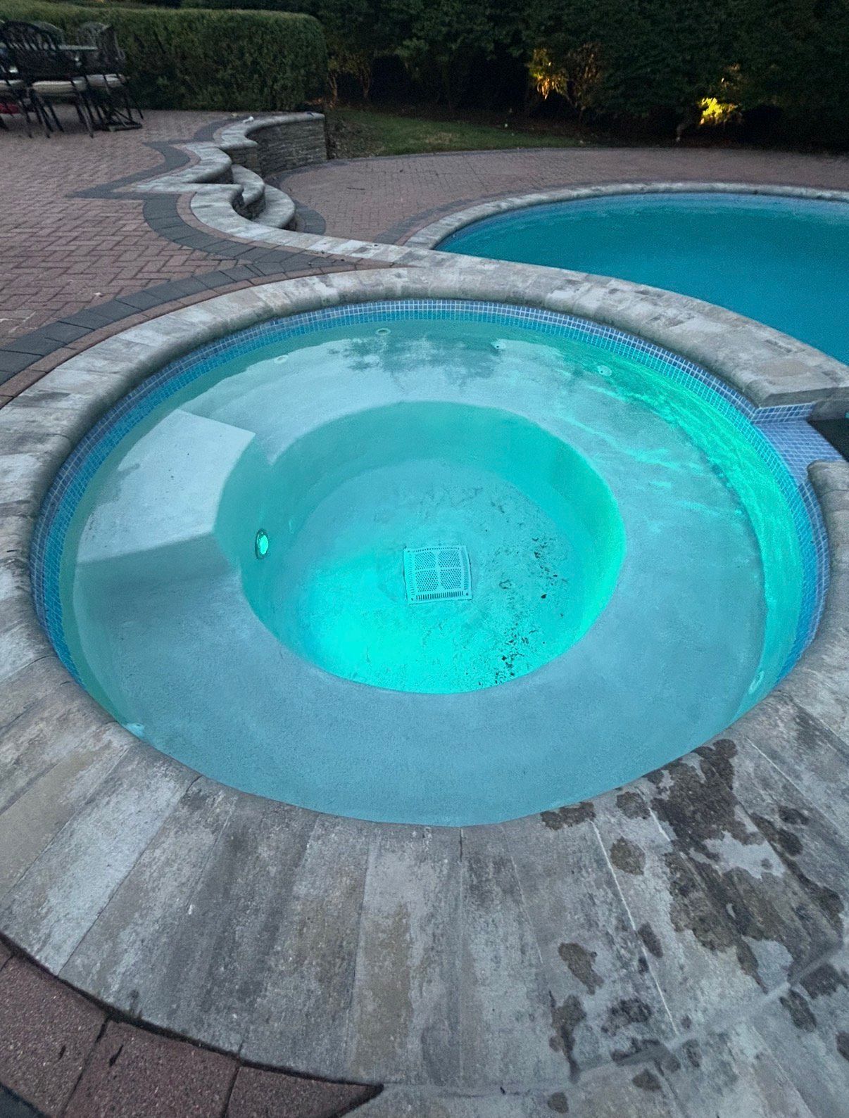 Circular hot tub with blue-green water surrounded by stone and a pool in the background.