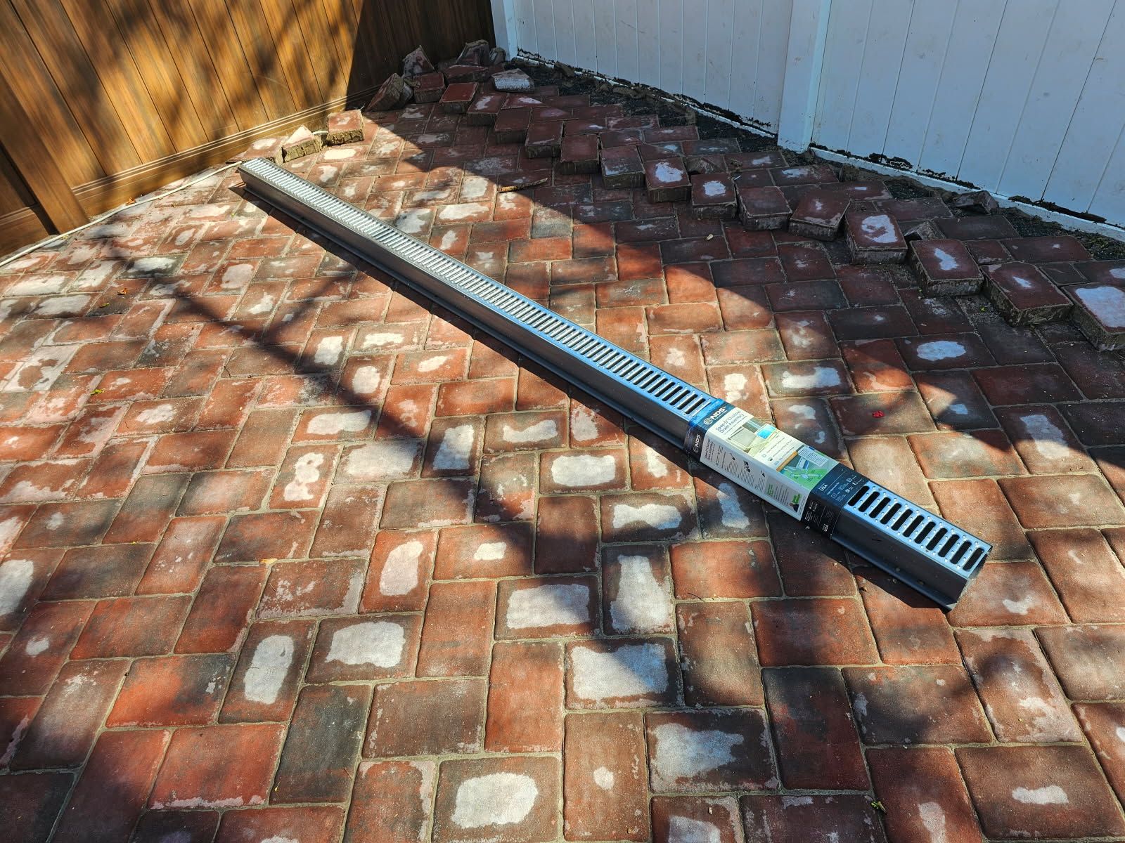 A long, black drainage channel on a red brick patio near a white wall and a wooden fence.