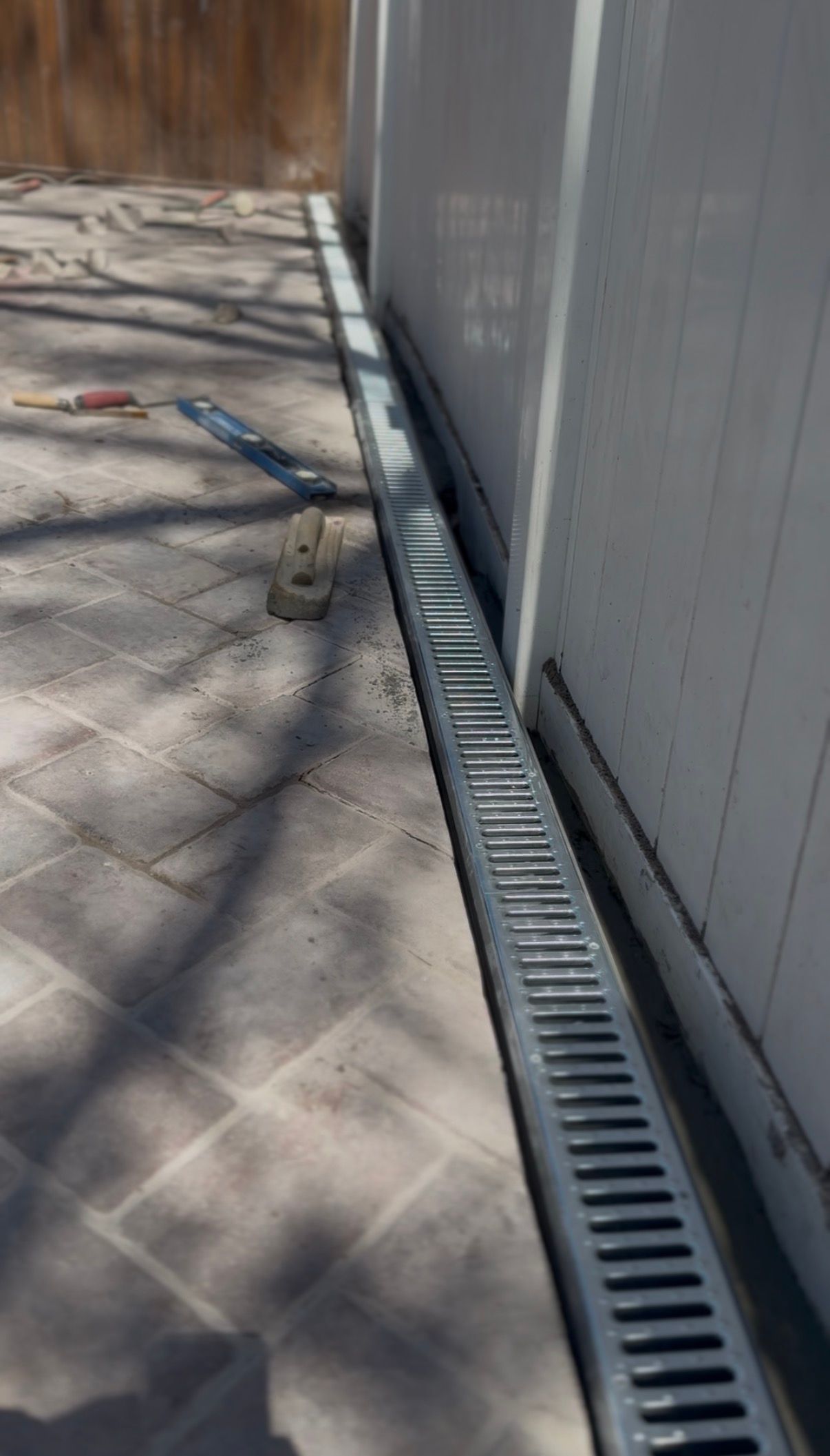 Grating drain along a white wall on a brick patio, casting shadows on a sunny day.