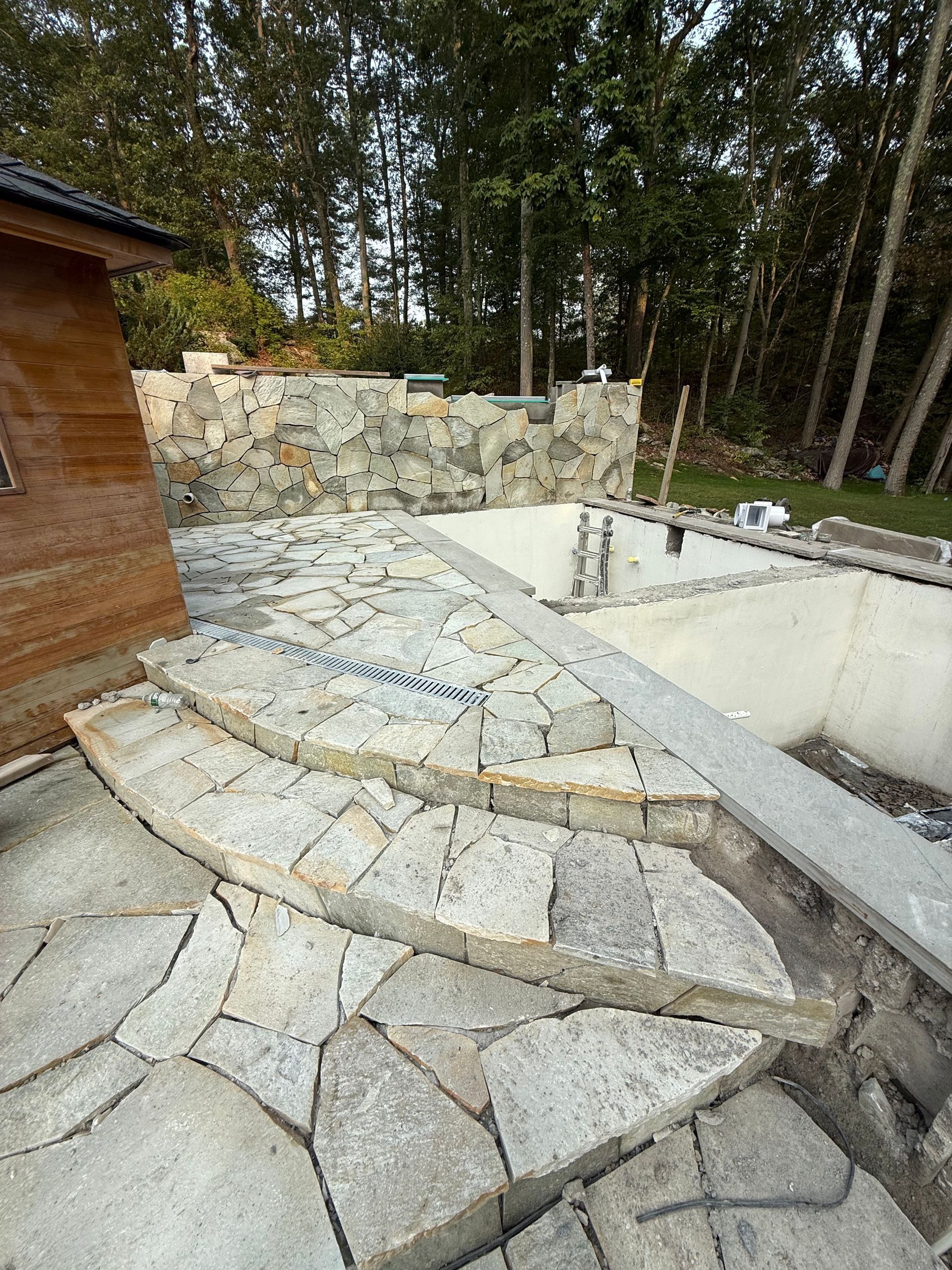 Stone steps leading to a partially built swimming pool, stone wall in the background, surrounded by trees.