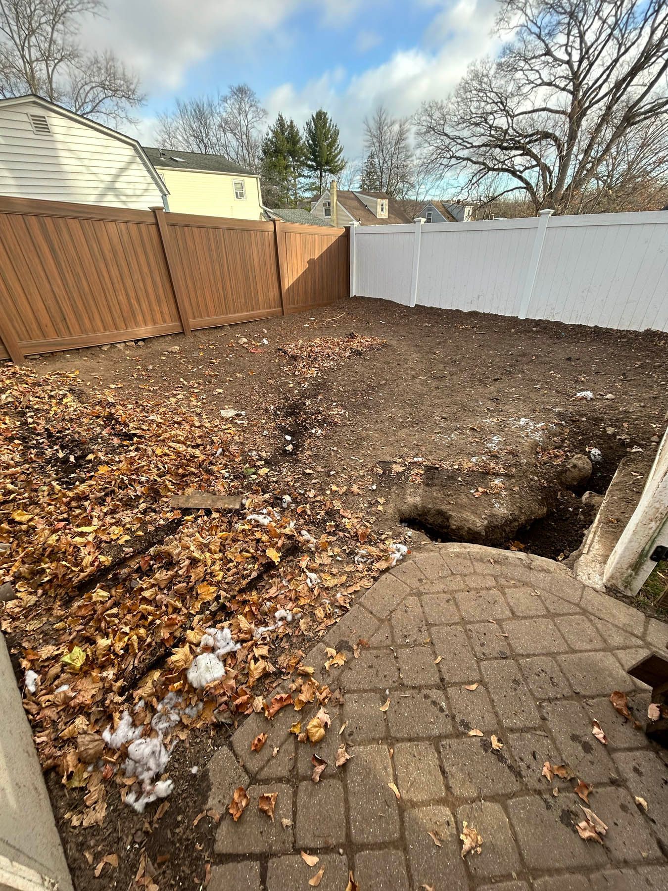Backyard with brown and white fences, bare soil, fallen leaves, and brick patio.