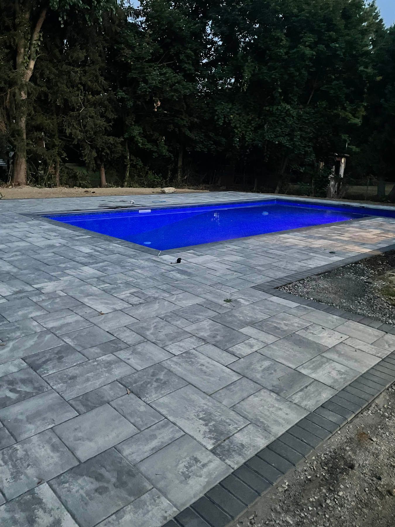 Blue illuminated rectangular pool surrounded by gray paving stones. Trees in the background.