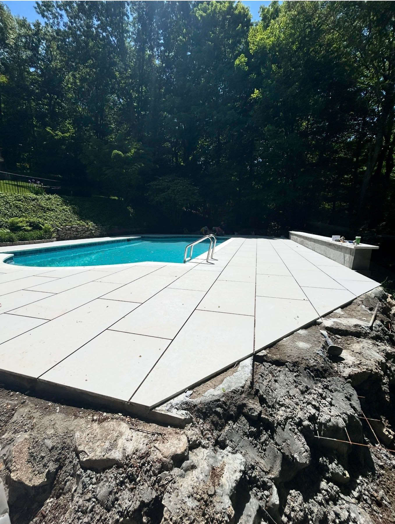 Pool deck with light-colored tiles, partially constructed, overlooking a blue pool. Trees in background.