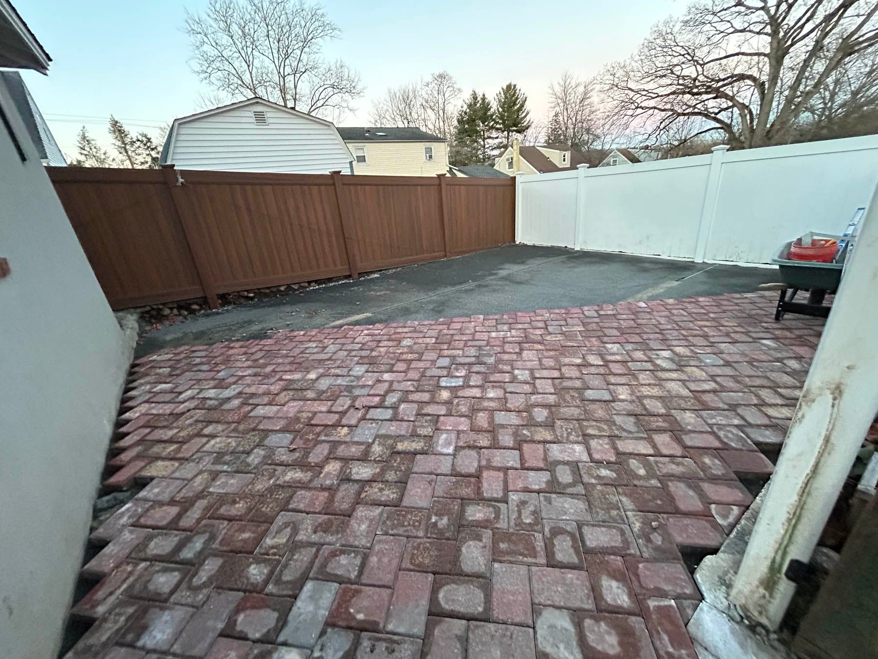 Brick patio leading to a driveway, bordered by brown and white fences.