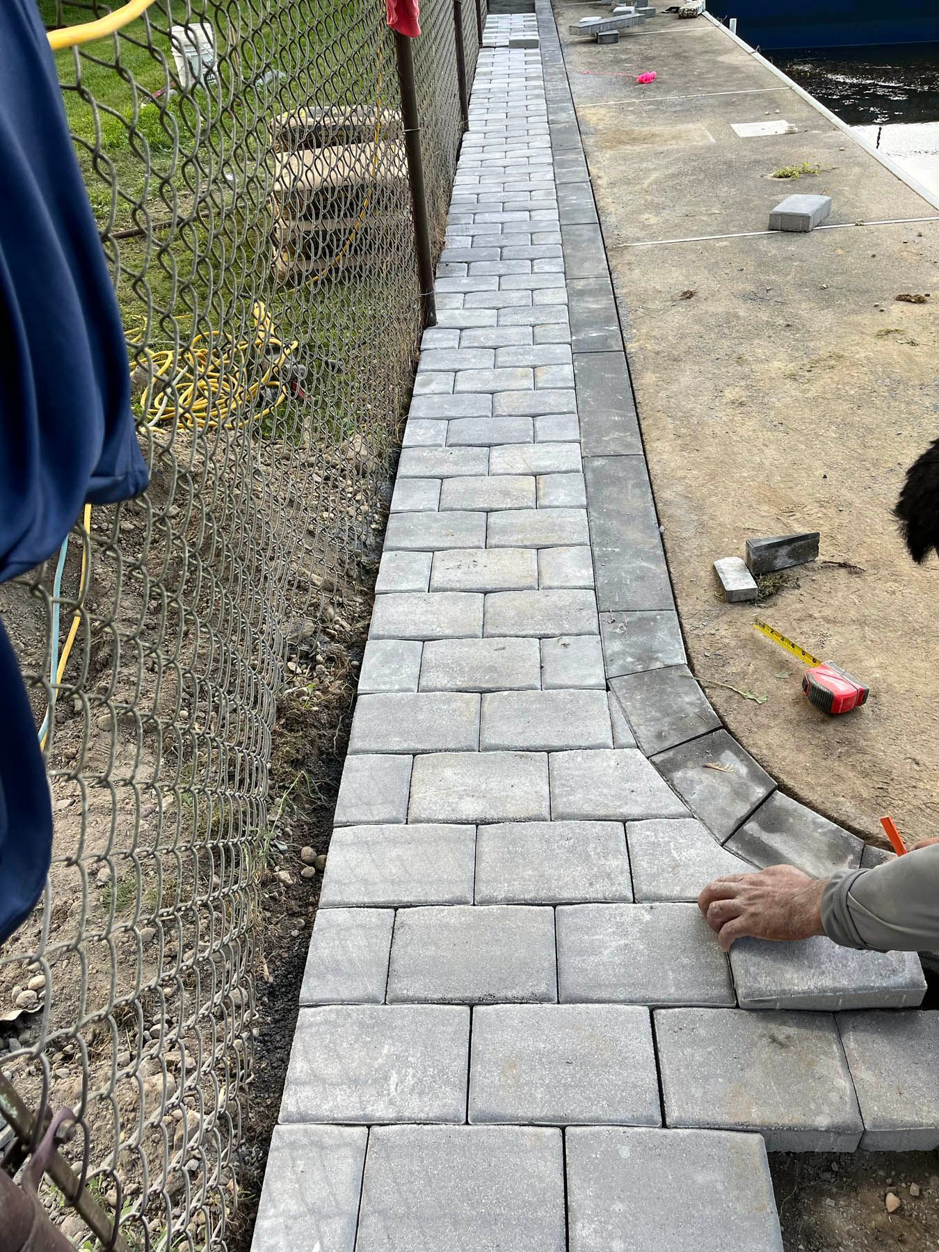 Installing gray pavers for a walkway next to a chain-link fence and concrete surface; a person lays a paver.