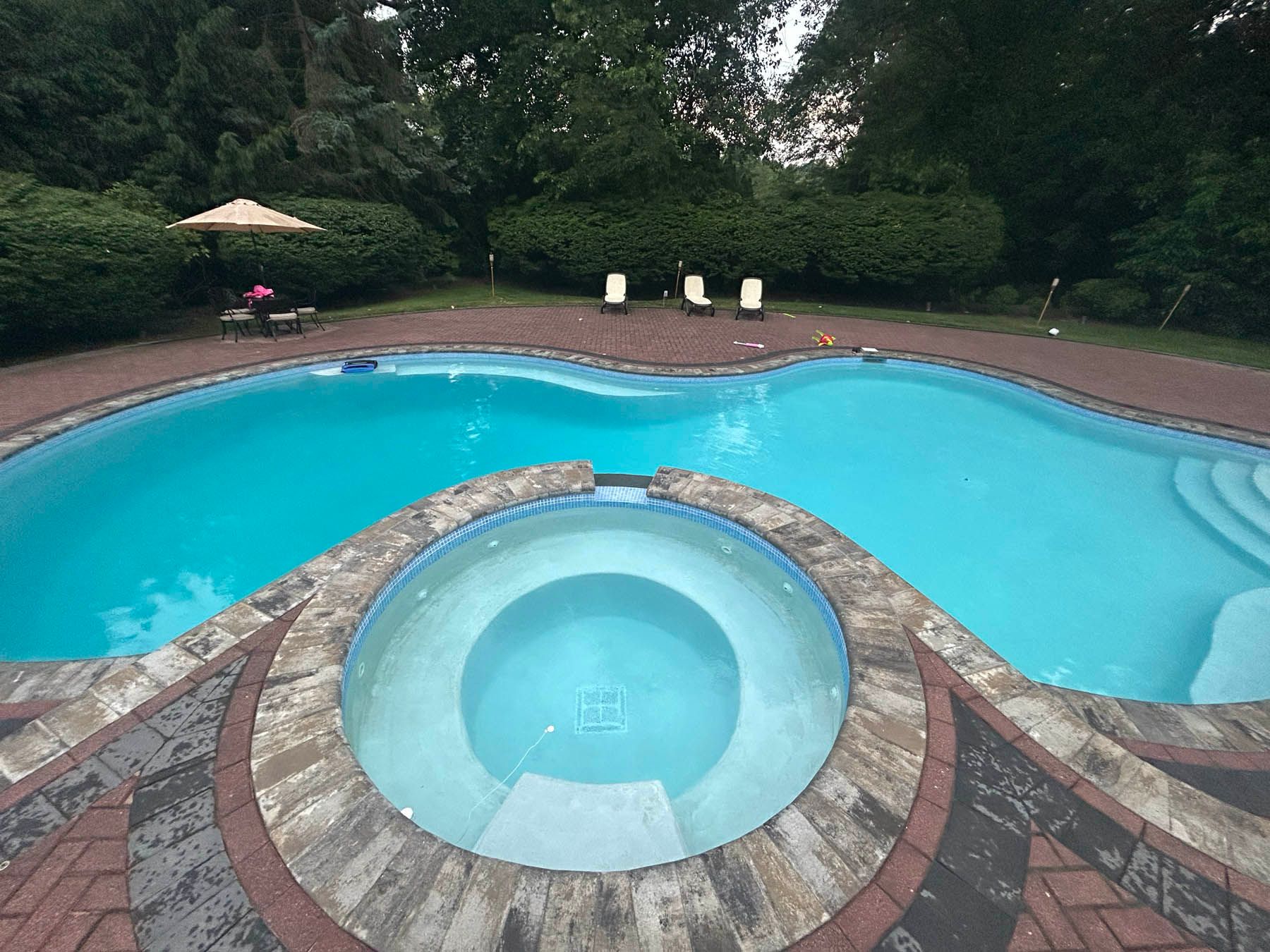 Pool with attached jacuzzi, brick border, lounge chairs, and umbrella in a lush outdoor setting.