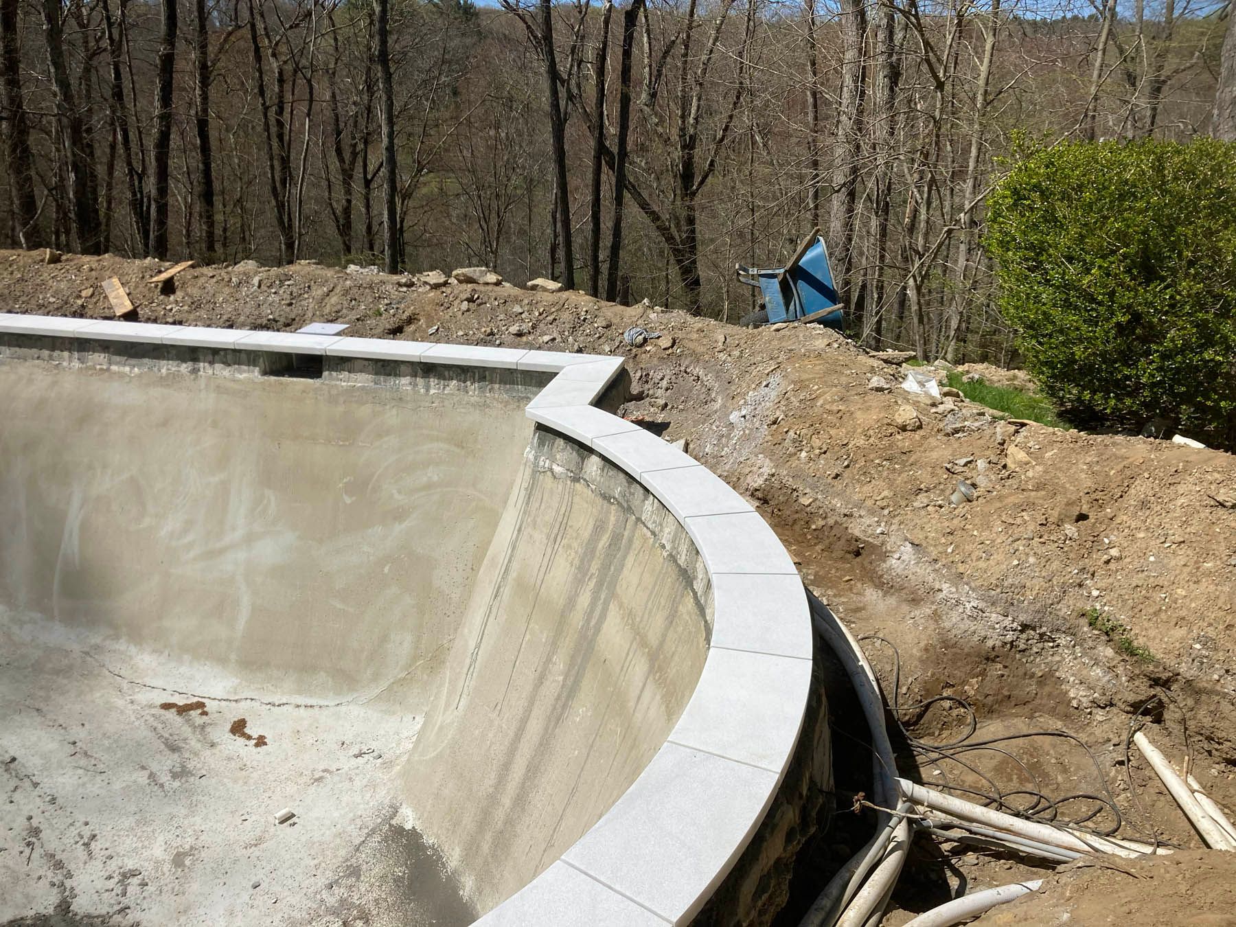 Unfinished swimming pool under construction, concrete walls with coping stones, surrounded by dirt and trees.