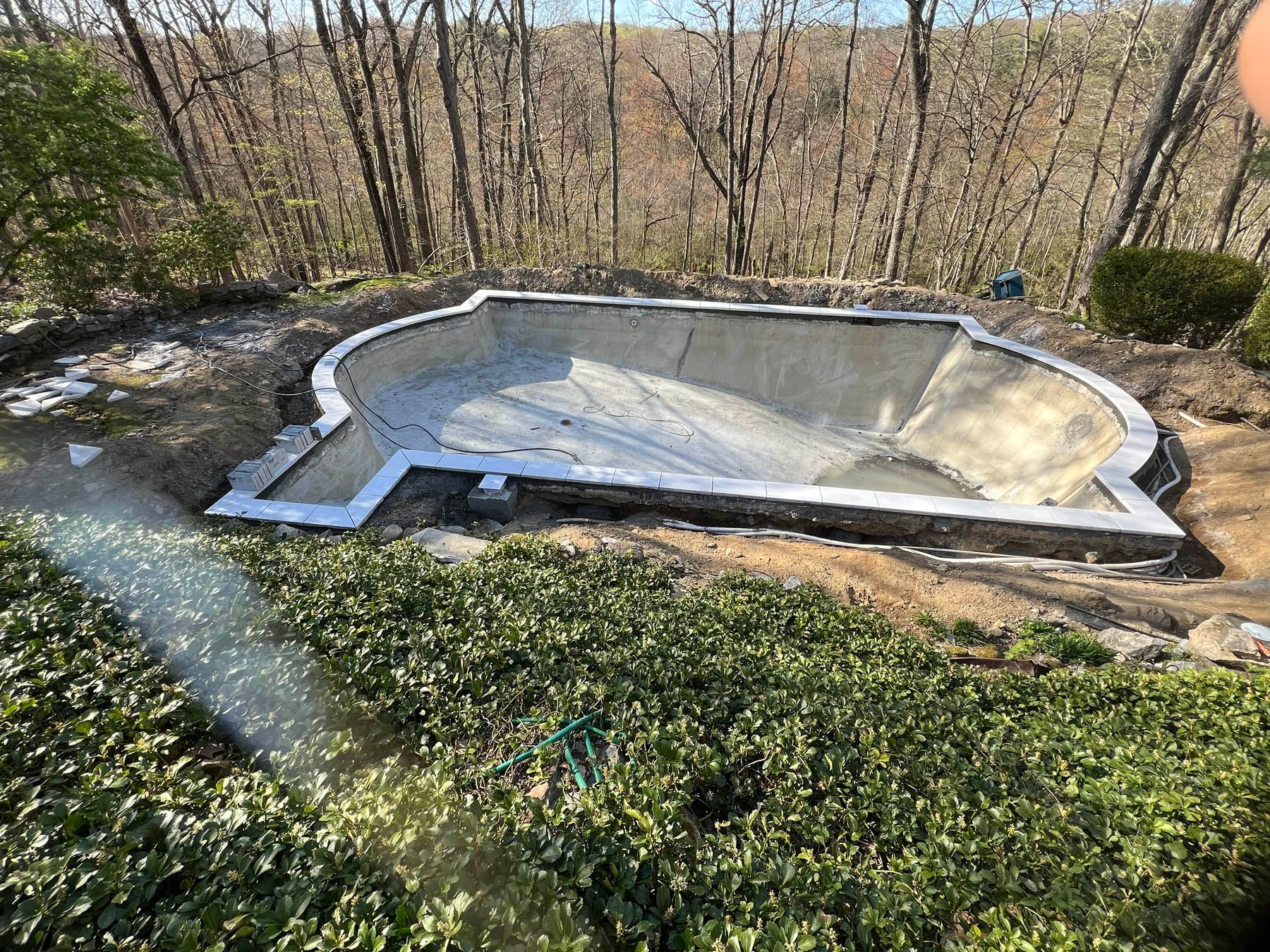 Construction of an empty, kidney-shaped swimming pool in a wooded backyard setting.