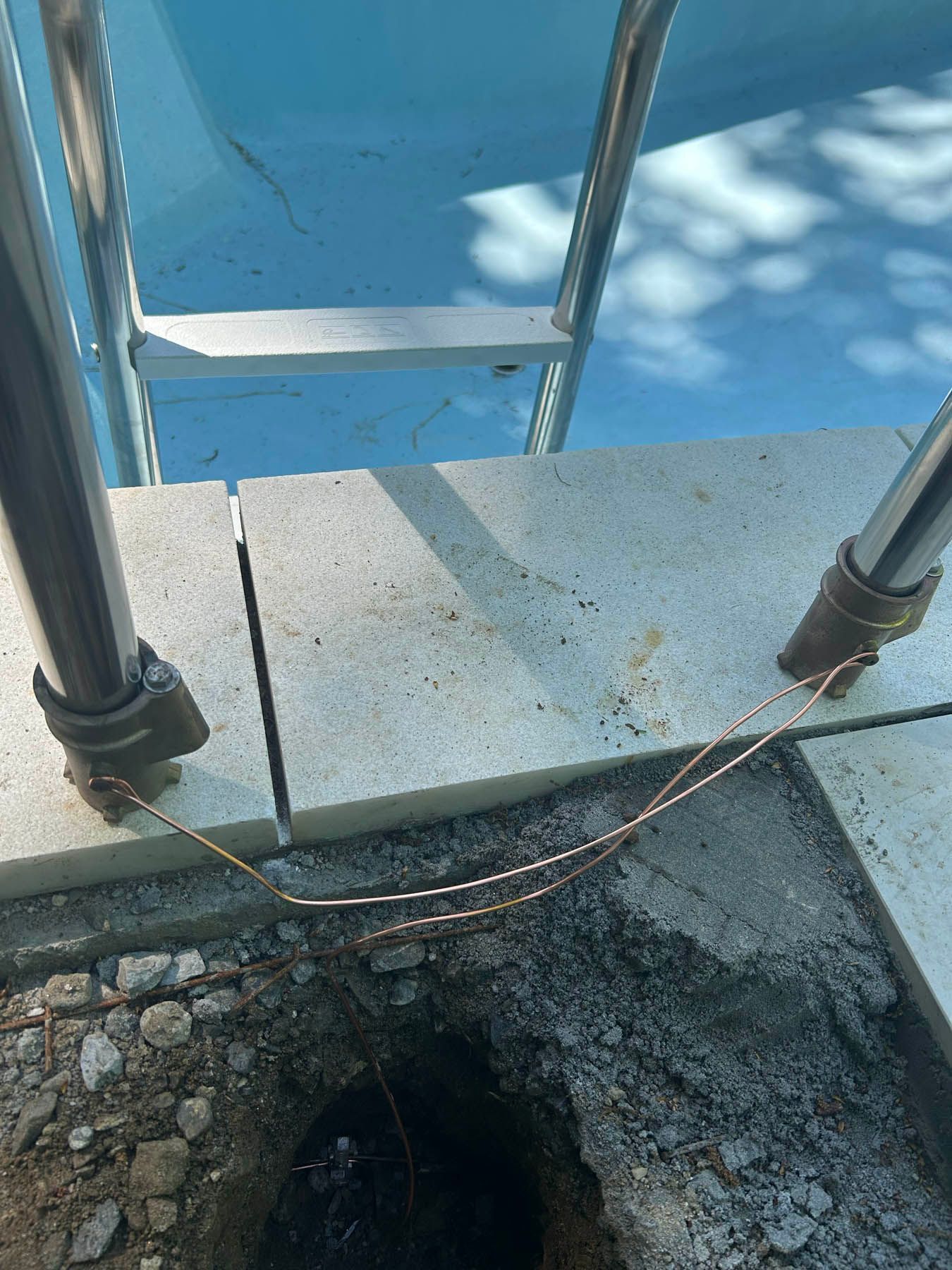 Pool ladder with exposed copper grounding wire near a hole in the concrete.