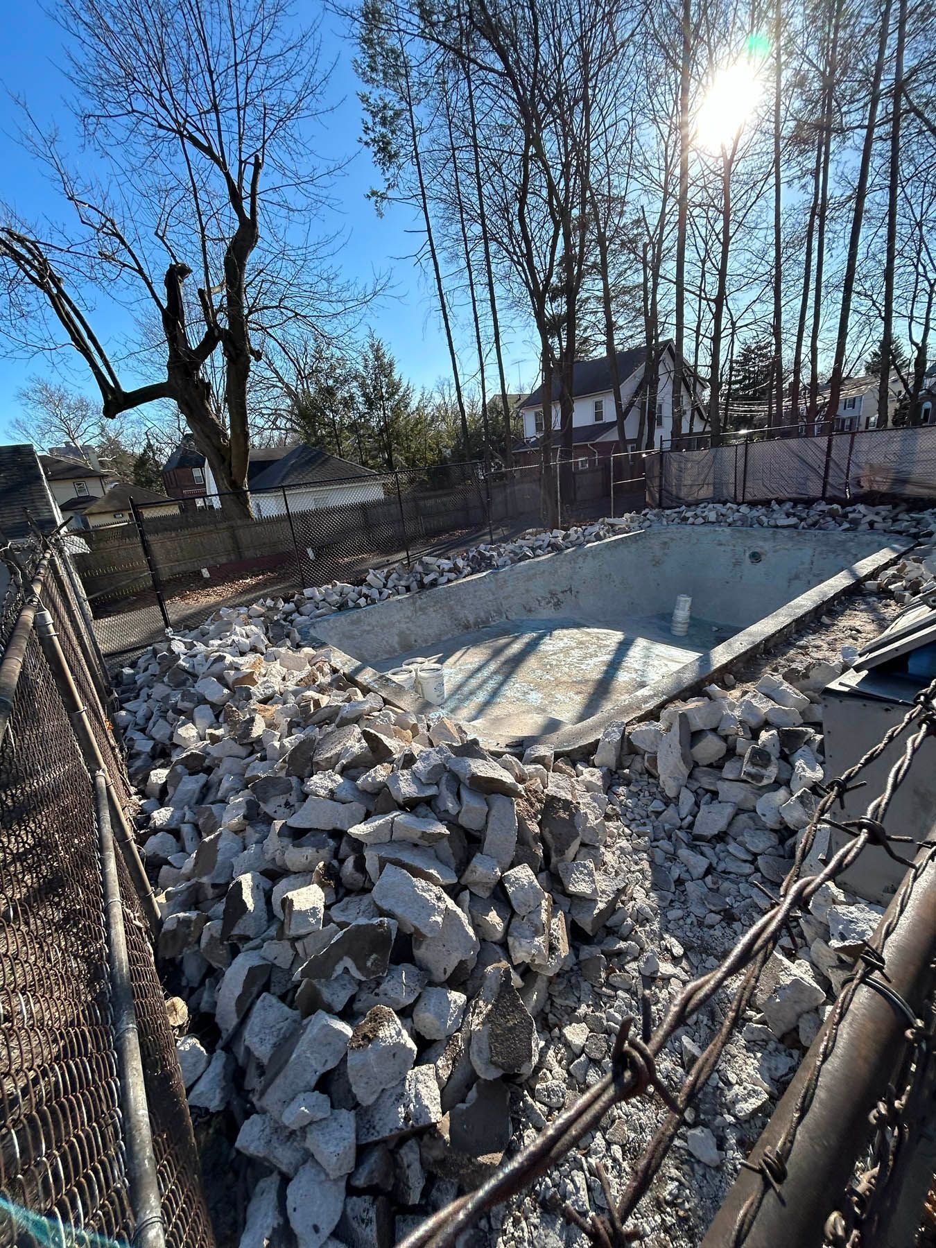 Pool demolition site: Concrete rubble, exposed pool shell, chain link fence, and sunlit sky.