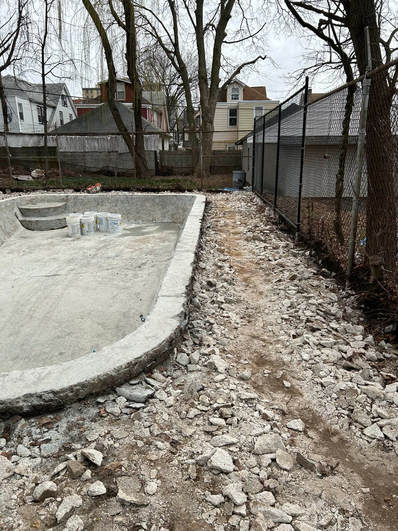 Concrete pool under construction, debris on ground, chain-link fence, trees, and houses.