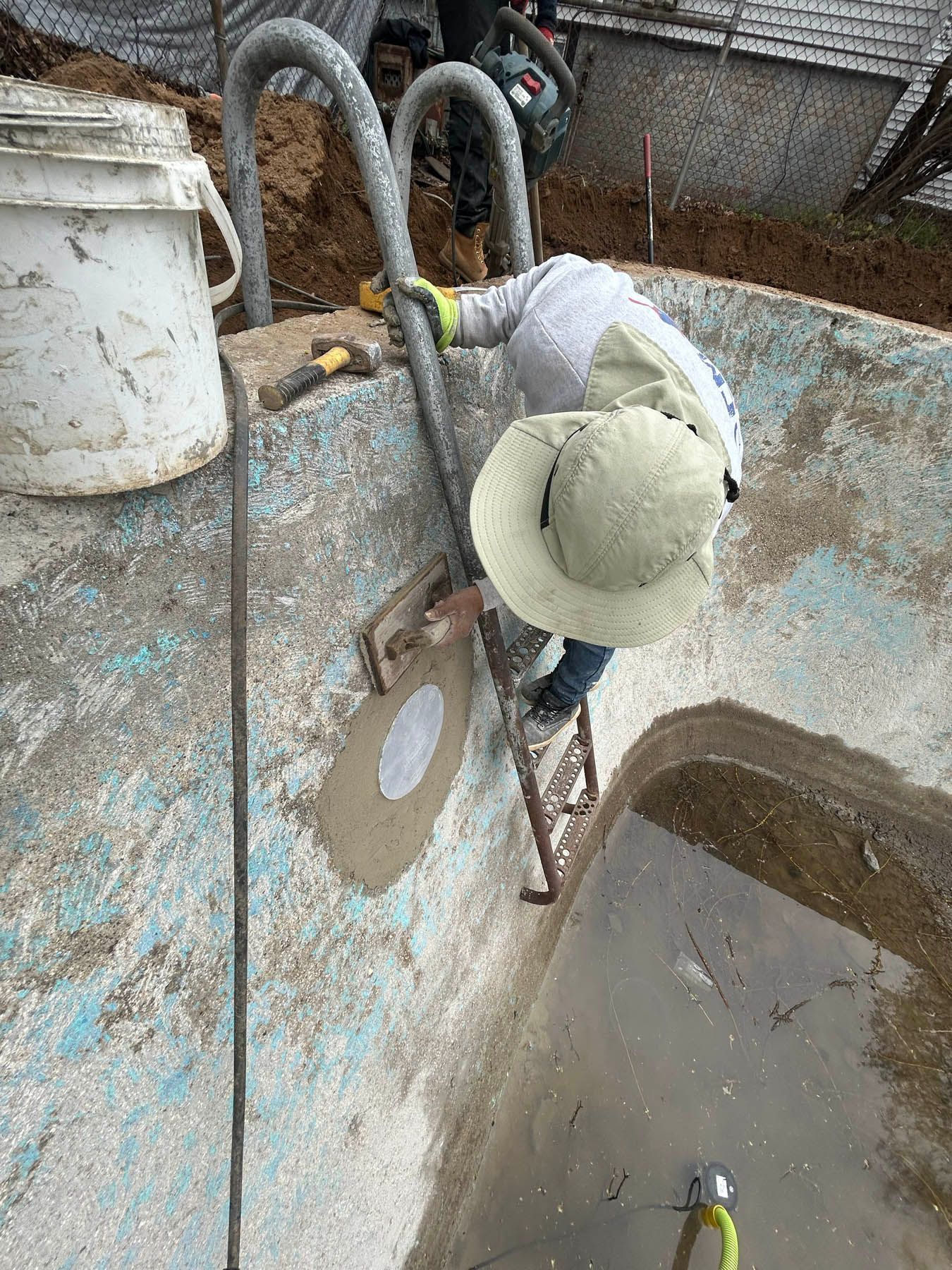 Person in work clothes climbs a pool ladder in a partially drained pool, bucket and tools nearby.