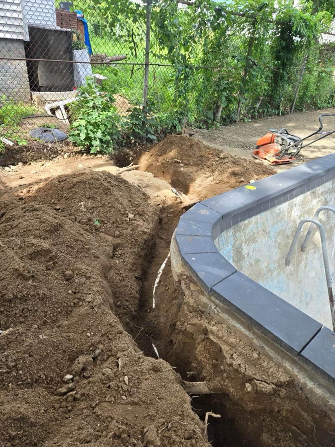 Trench dug near a pool, exposing pipes. Dirt pile and small compactor visible.
