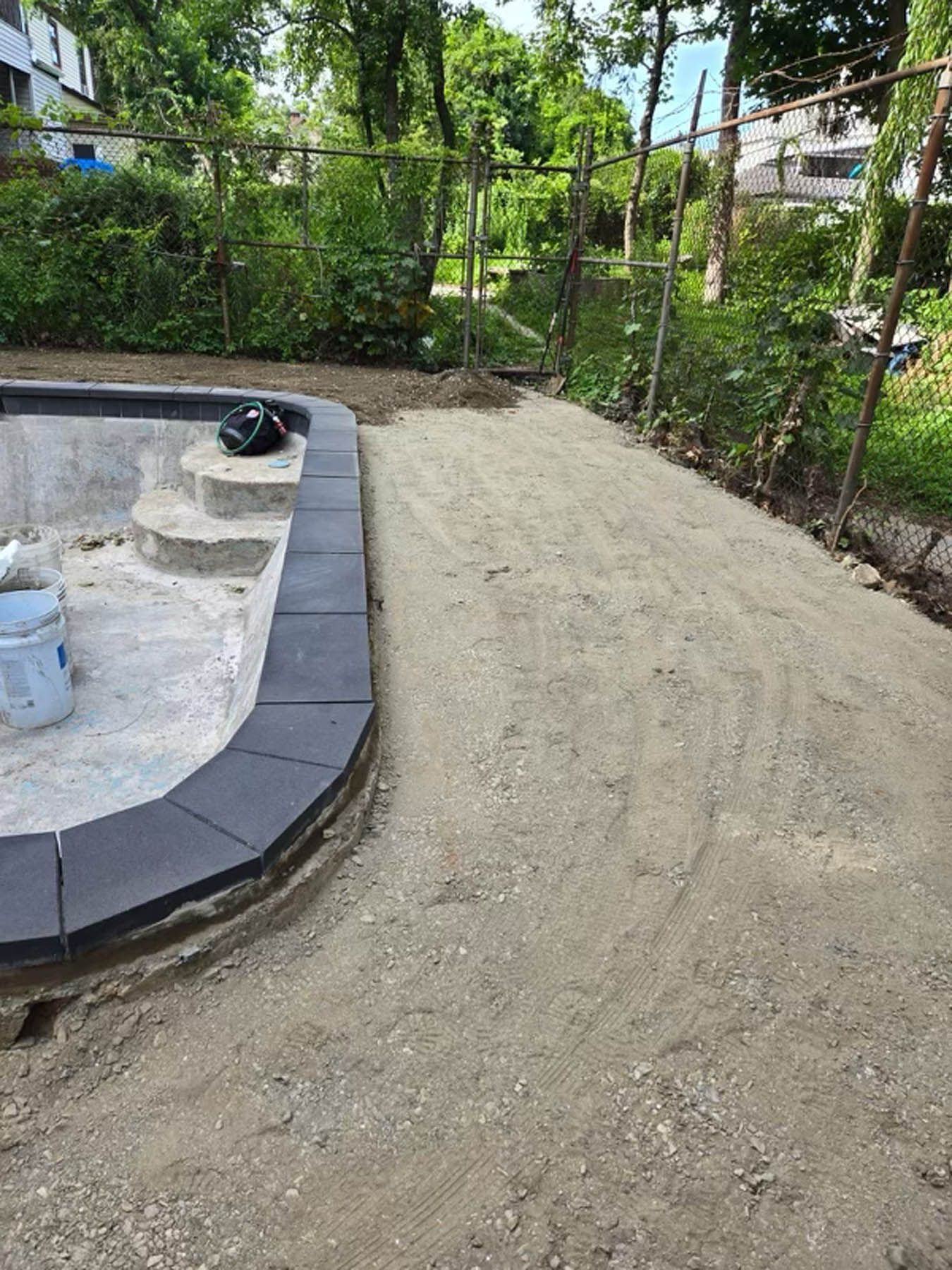 Poolside construction: concrete pool with gray pavers, unfinished gravel path, and chain-link fence.