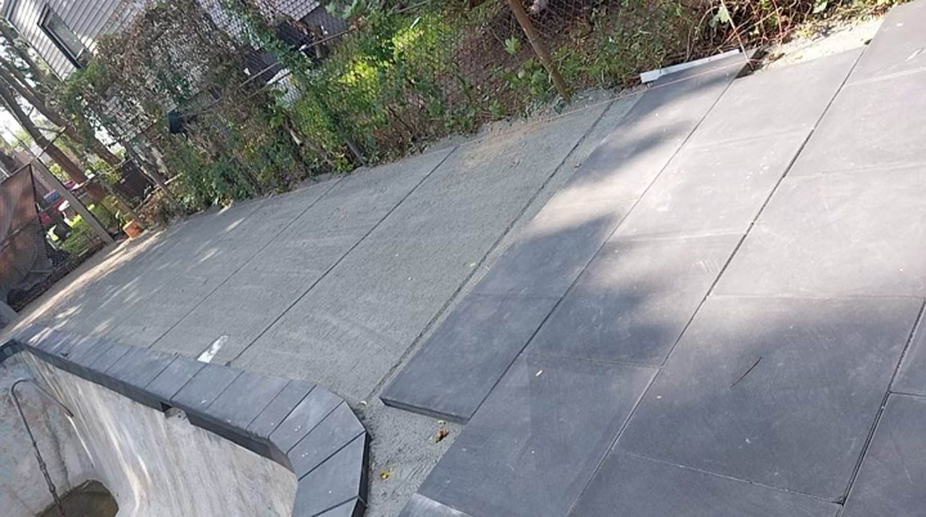 Gray stone pavers laid in rows, adjacent to a curved border. Background features greenery and a partial fence.