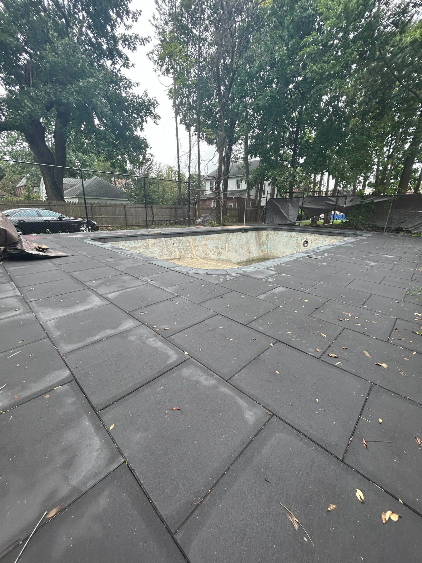 Empty swimming pool surrounded by dark gray rubber tiles and trees on a cloudy day.
