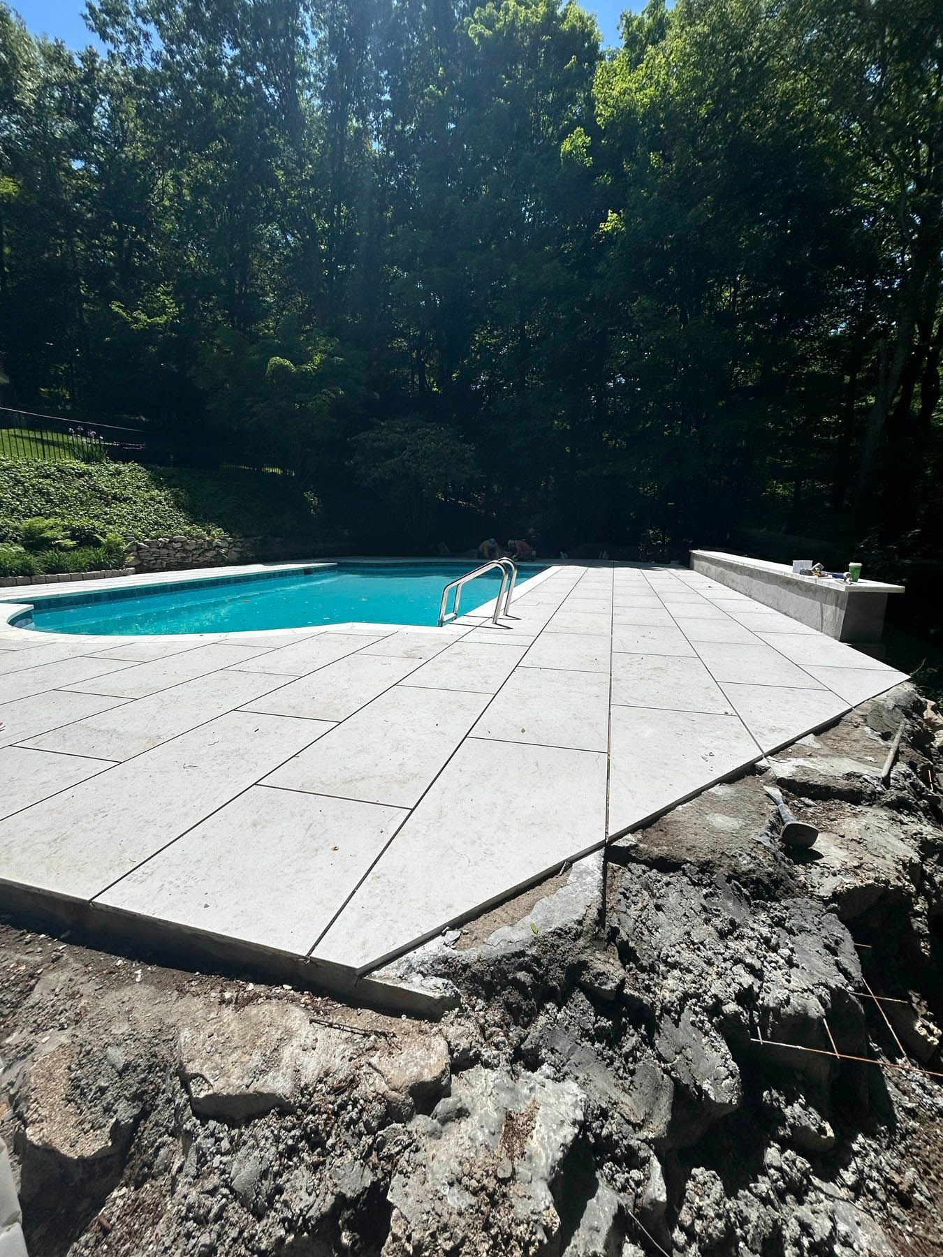 Pool with light-colored stone patio, surrounded by rocks and trees. Blue water reflects sunlight.