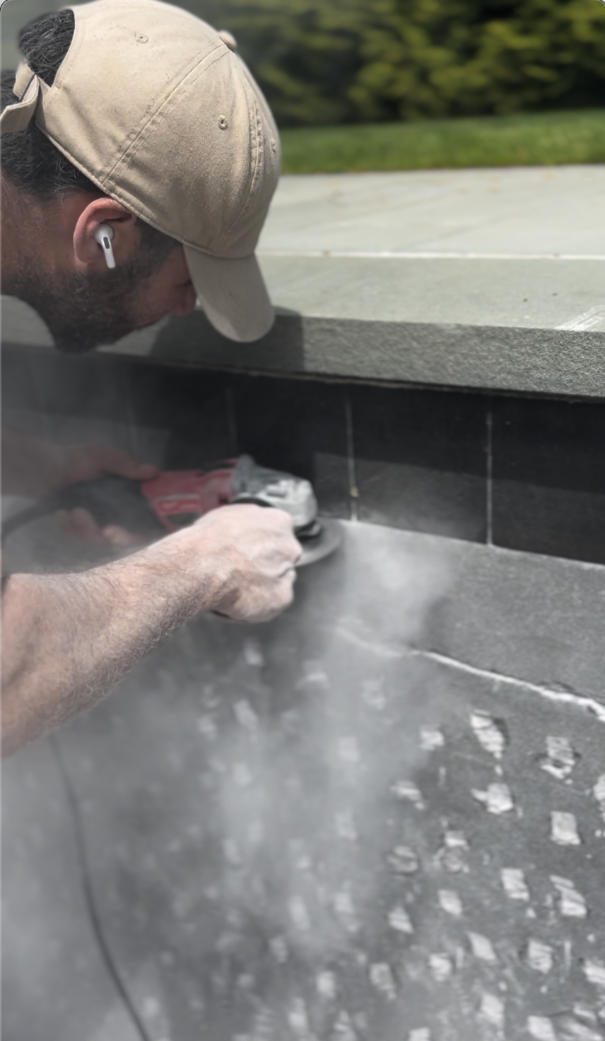 Man using a grinder on a stone wall, creating dust; outdoors.