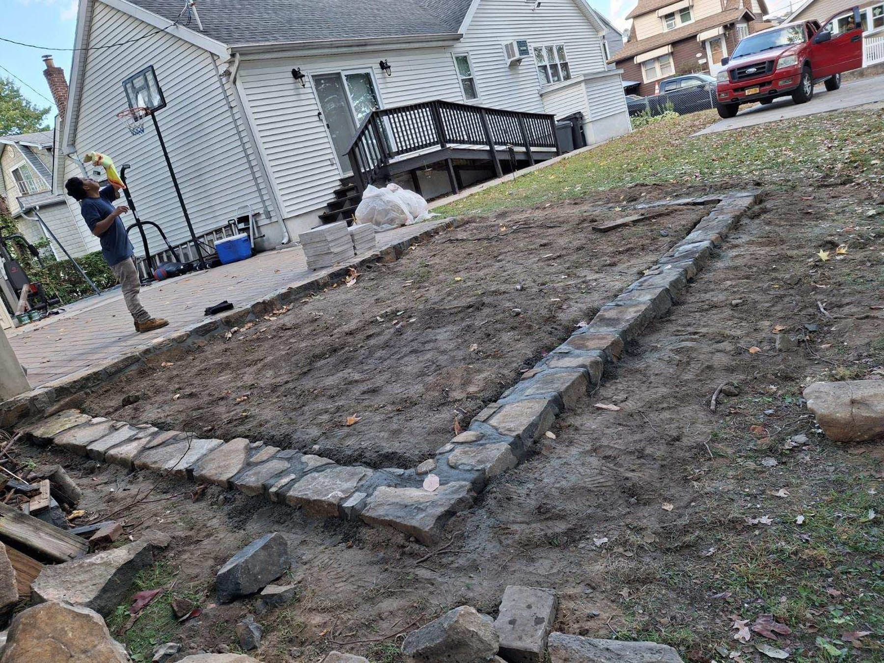 A person building a stone-bordered garden bed on a sloped yard.