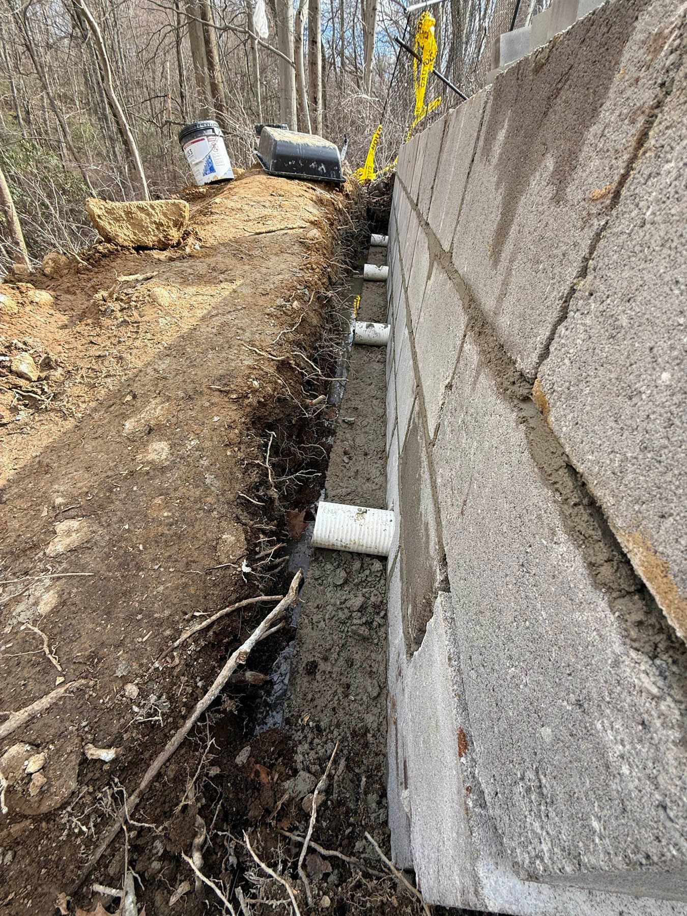 Trench with white pipes next to a gray cinder block wall. Dirt, debris, and buckets in the background.