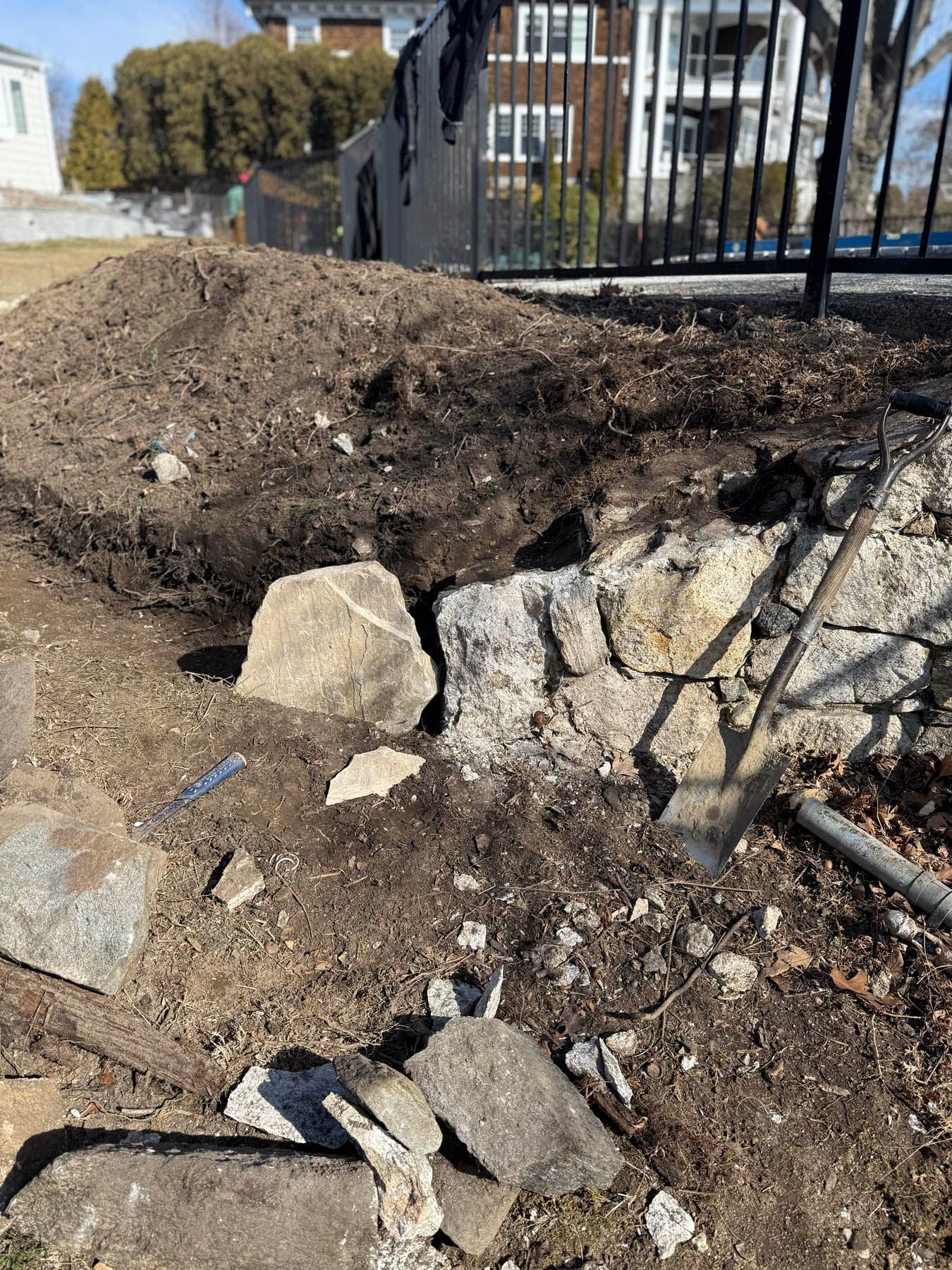 A rock wall being excavated; dirt, rocks, and a shovel are visible. Fence and trees in background.