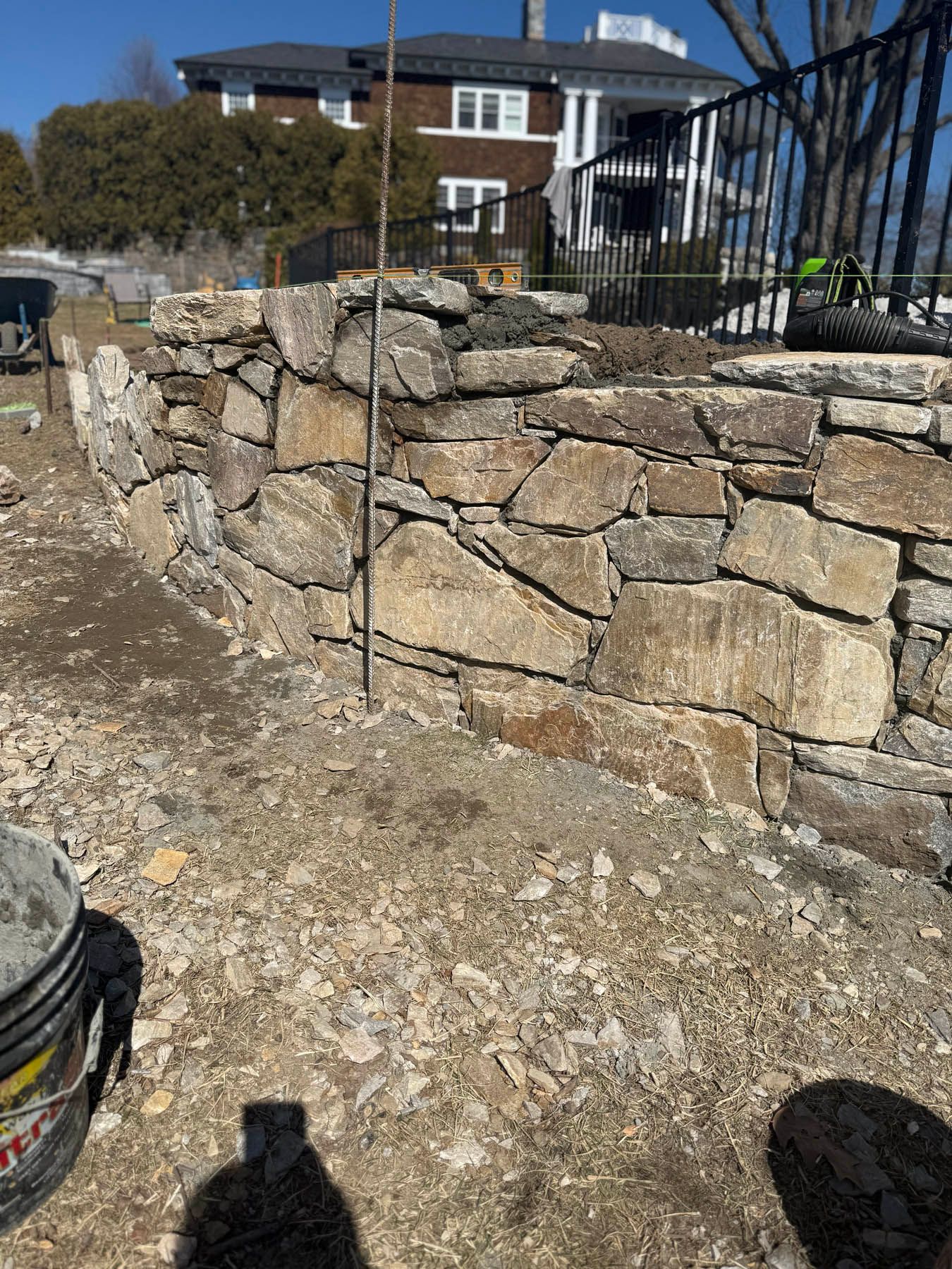 Stone wall being built outdoors, with construction materials and house in the background.