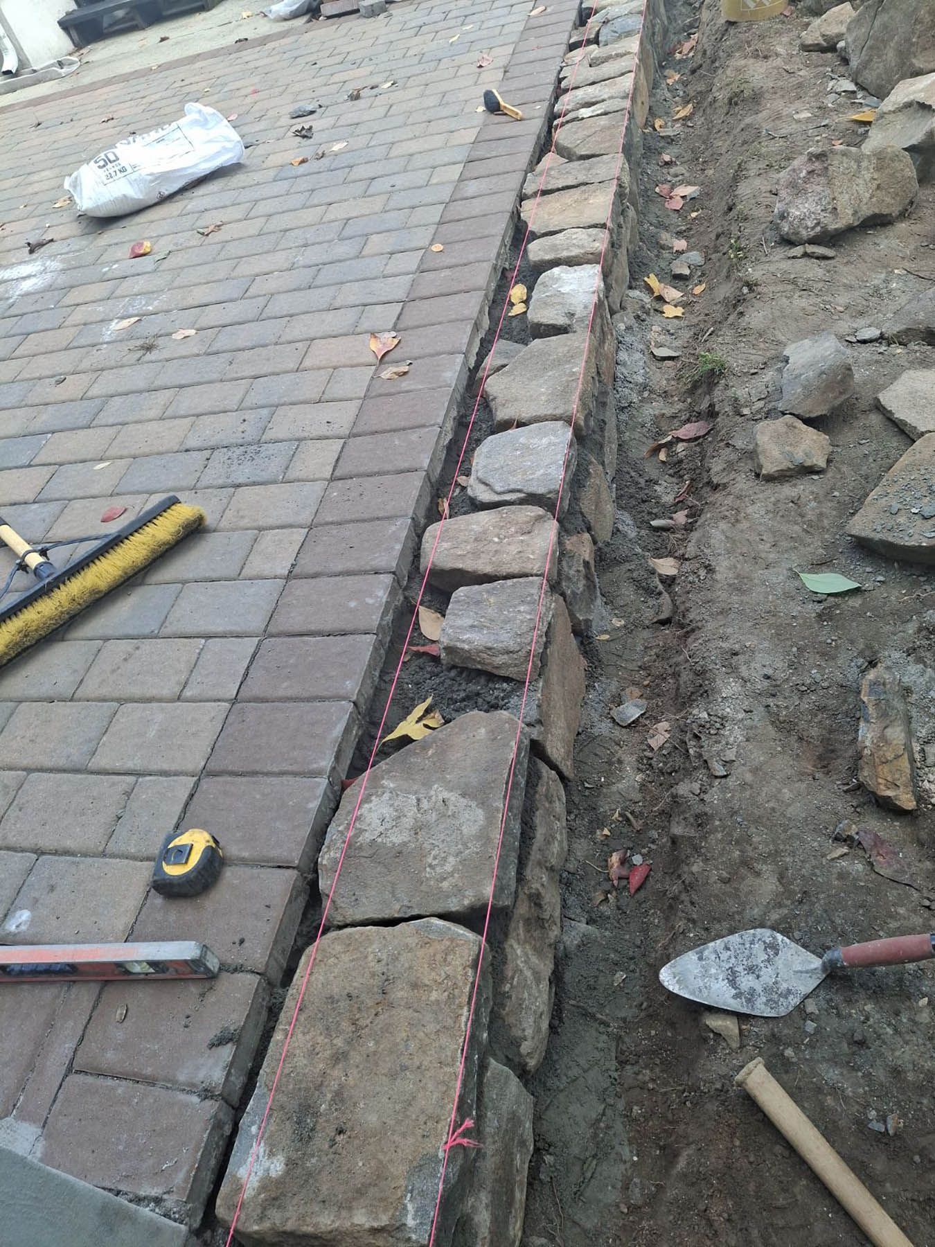 Stone border being installed along a brick paver walkway. Tools and materials are nearby.
