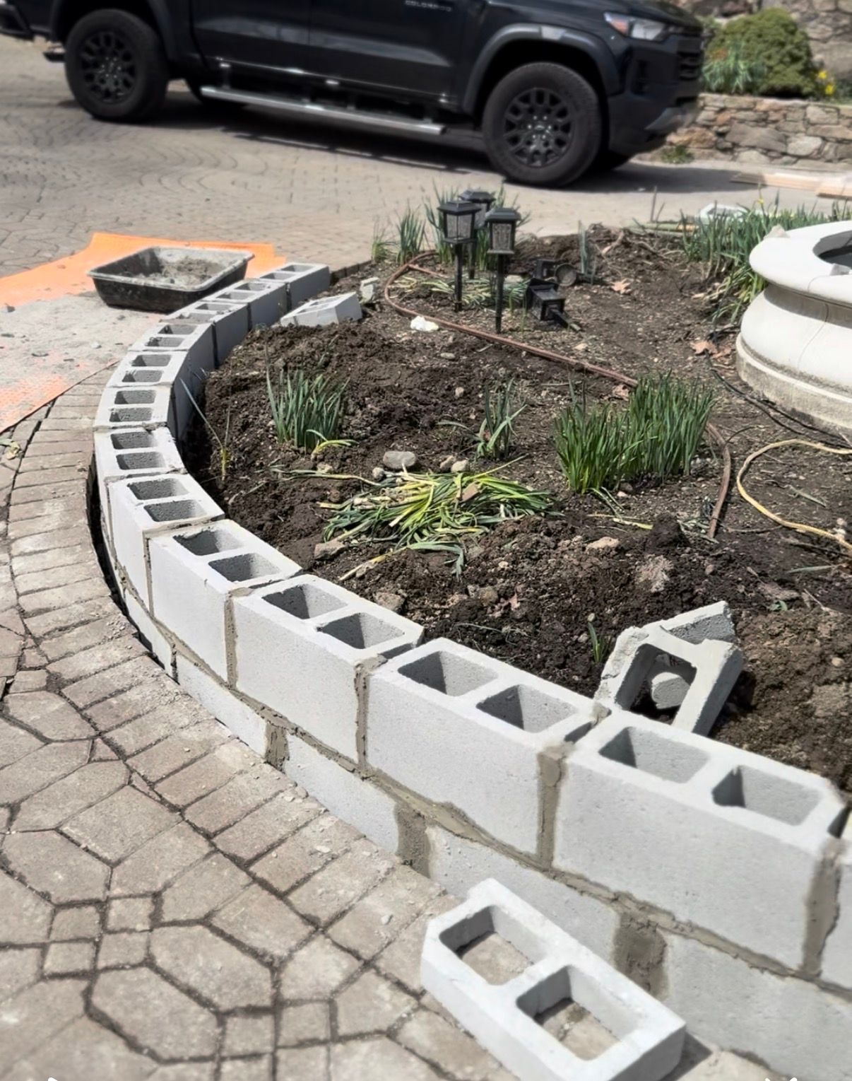 Curved cinder block retaining wall under construction in a garden bed with a vehicle in the background.