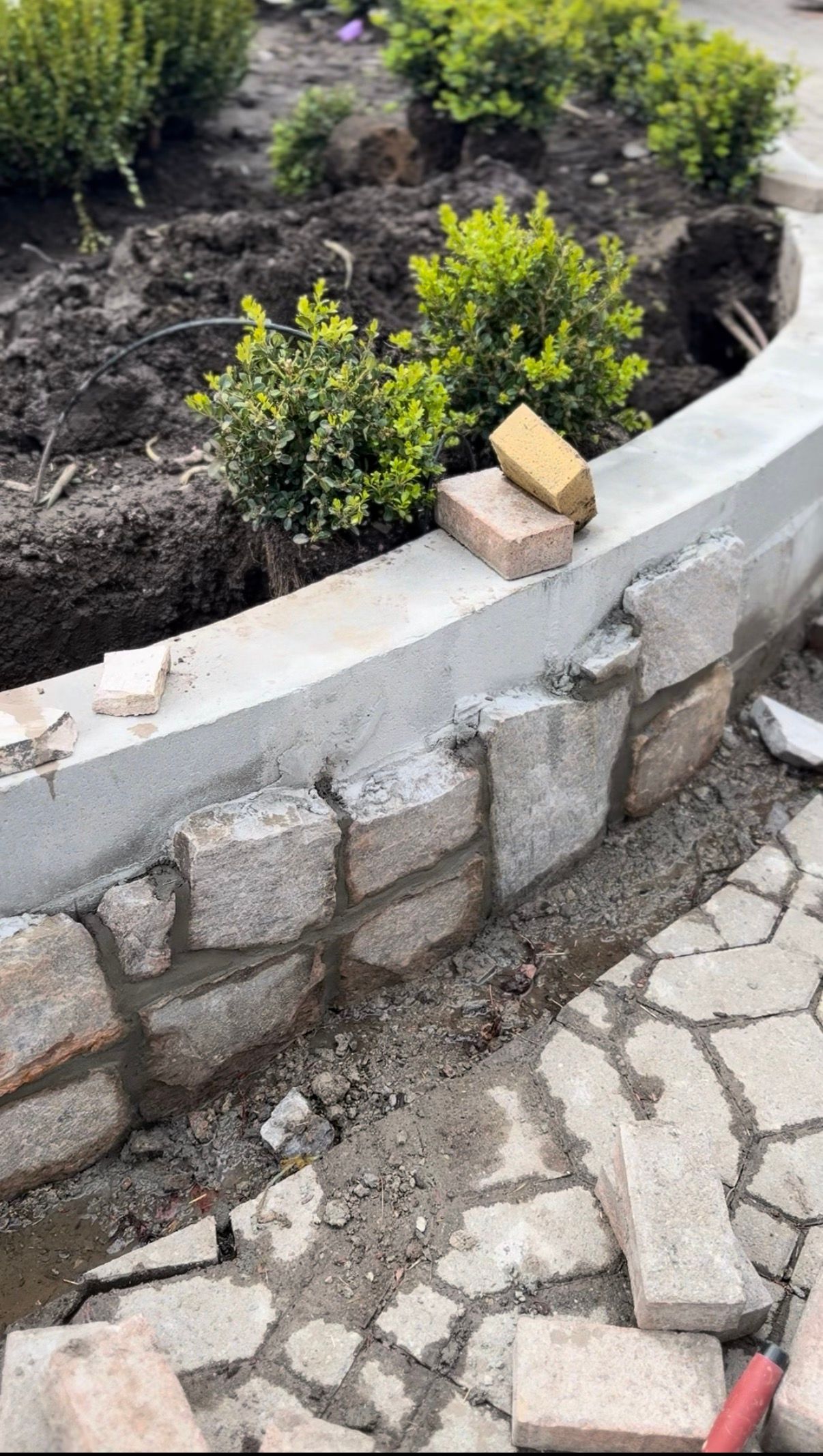 Brick planter under construction with plants in the background.