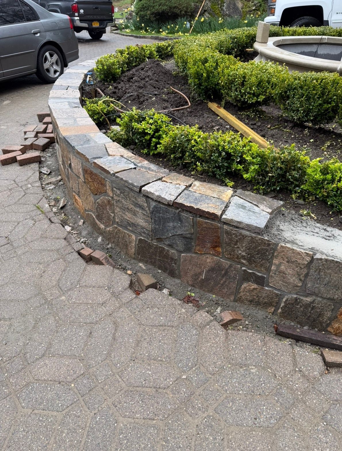Stone retaining wall with plants, part of a garden bed in front of a house, brick edging.