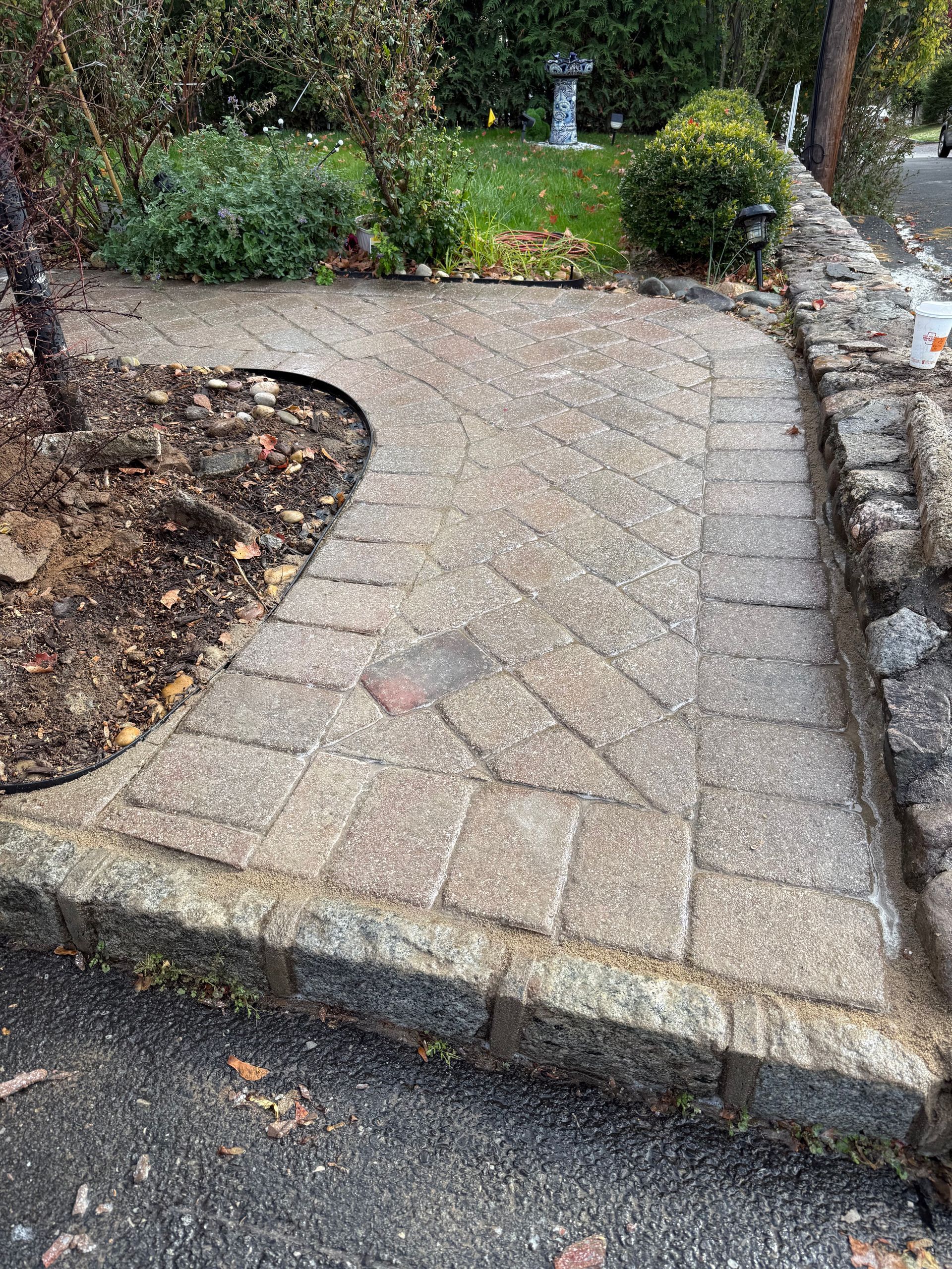 Stone pathway curving through a garden, bordered by a low stone wall and asphalt.