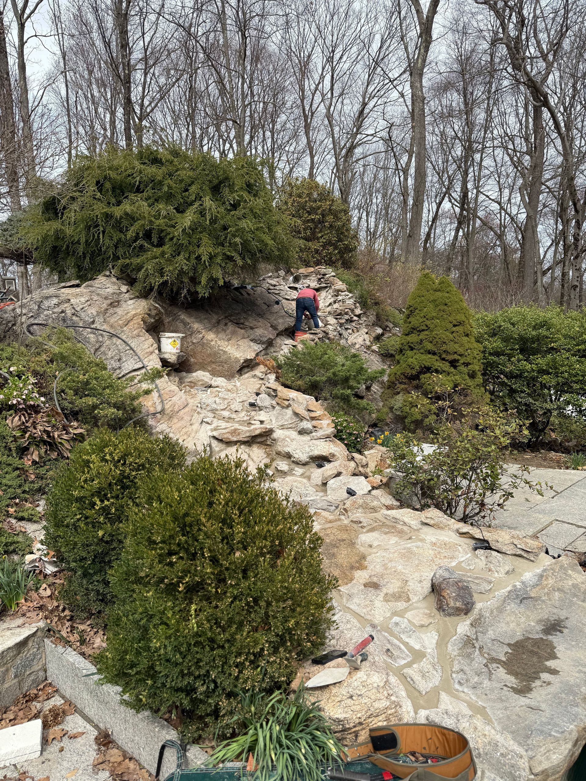 Rocky garden slope with green bushes and a person working; trees in the background.