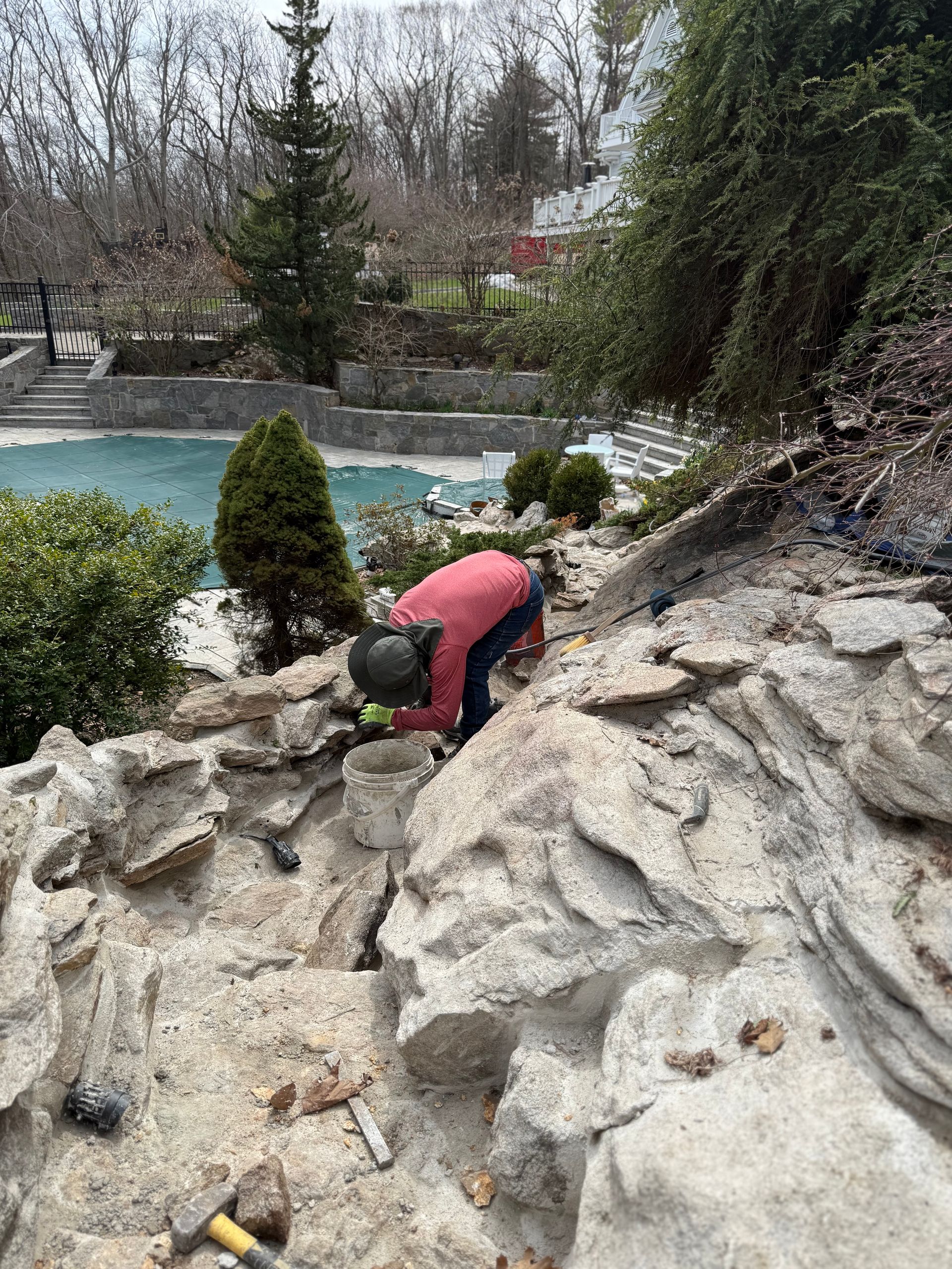 Person working on stone structure near a pool, using tools to place stones.