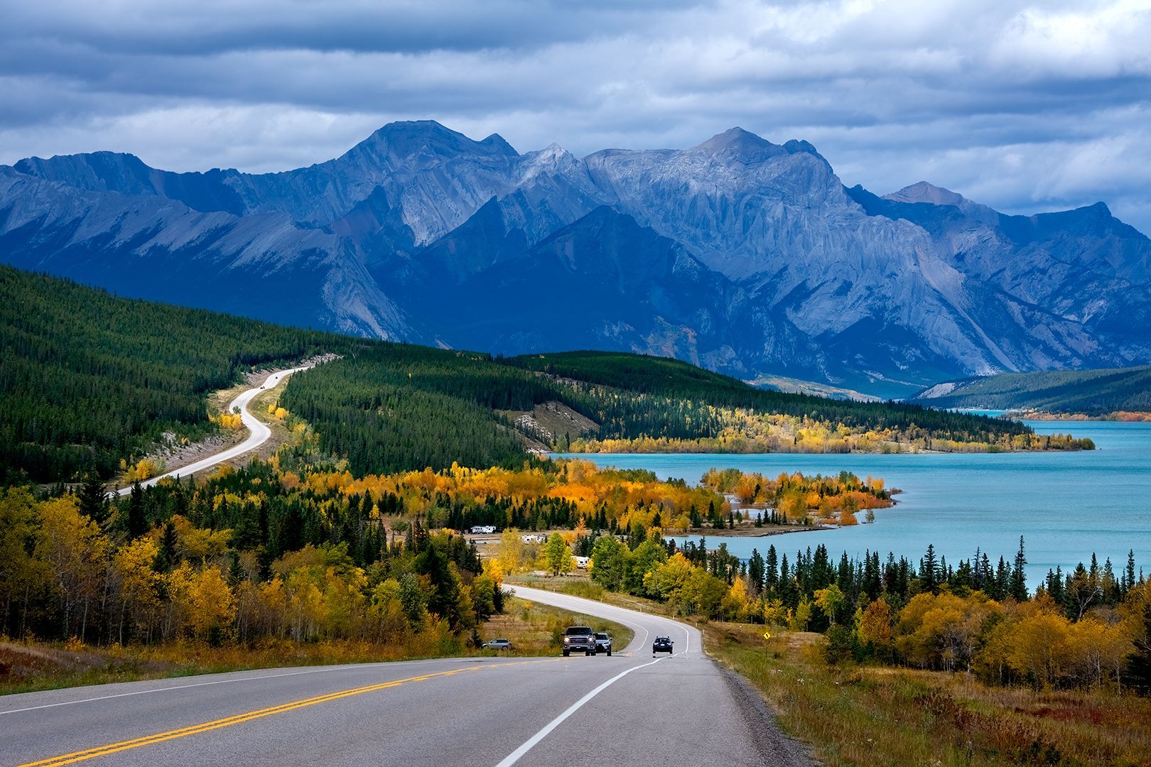 Winding road through autumn trees, turquoise lake, and towering blue mountains under a cloudy sky.